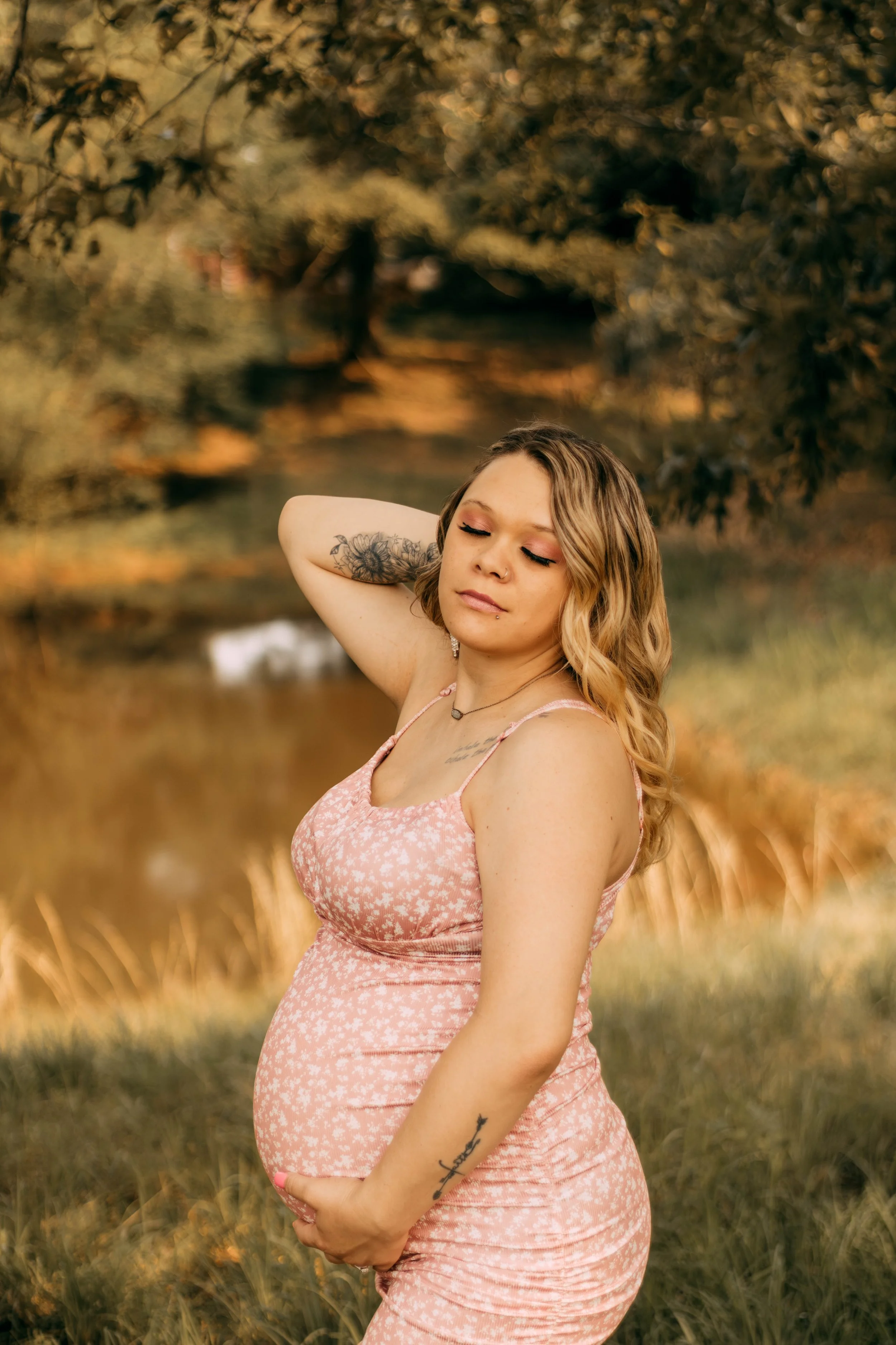 A pregnant woman in a pink floral dress outdoors near a pond, with her eyes closed, one hand on her neck, and the other resting on her belly, during late afternoon.