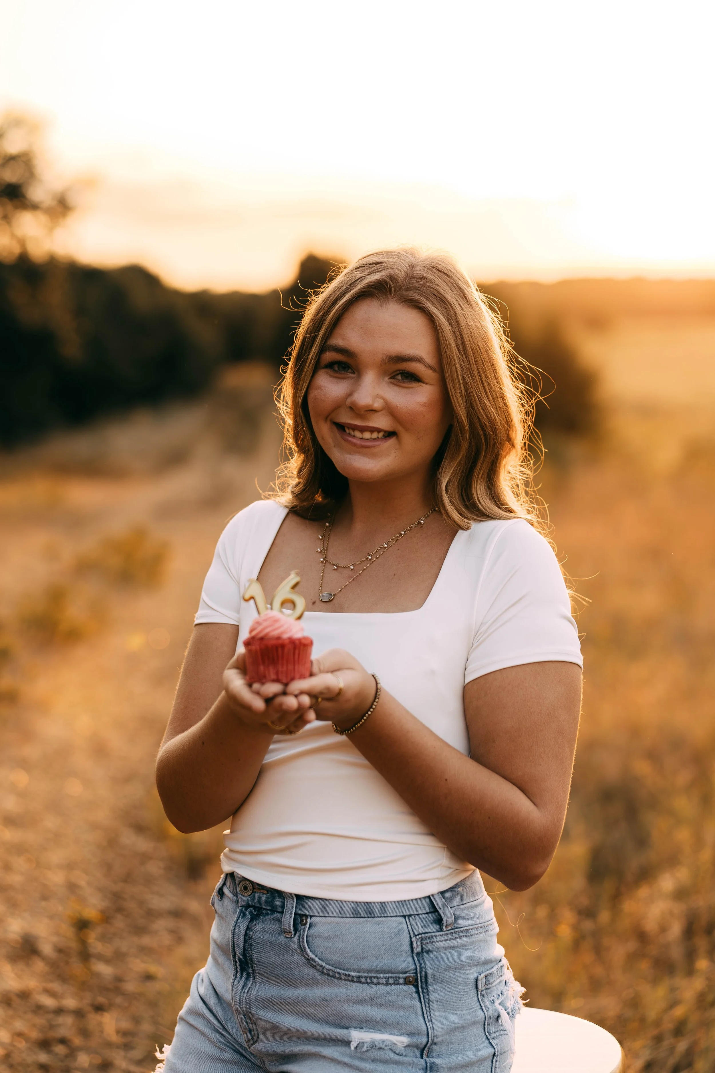 A young woman outdoors holding a cupcake with a birthday candle shaped like the number 16, during sunset.