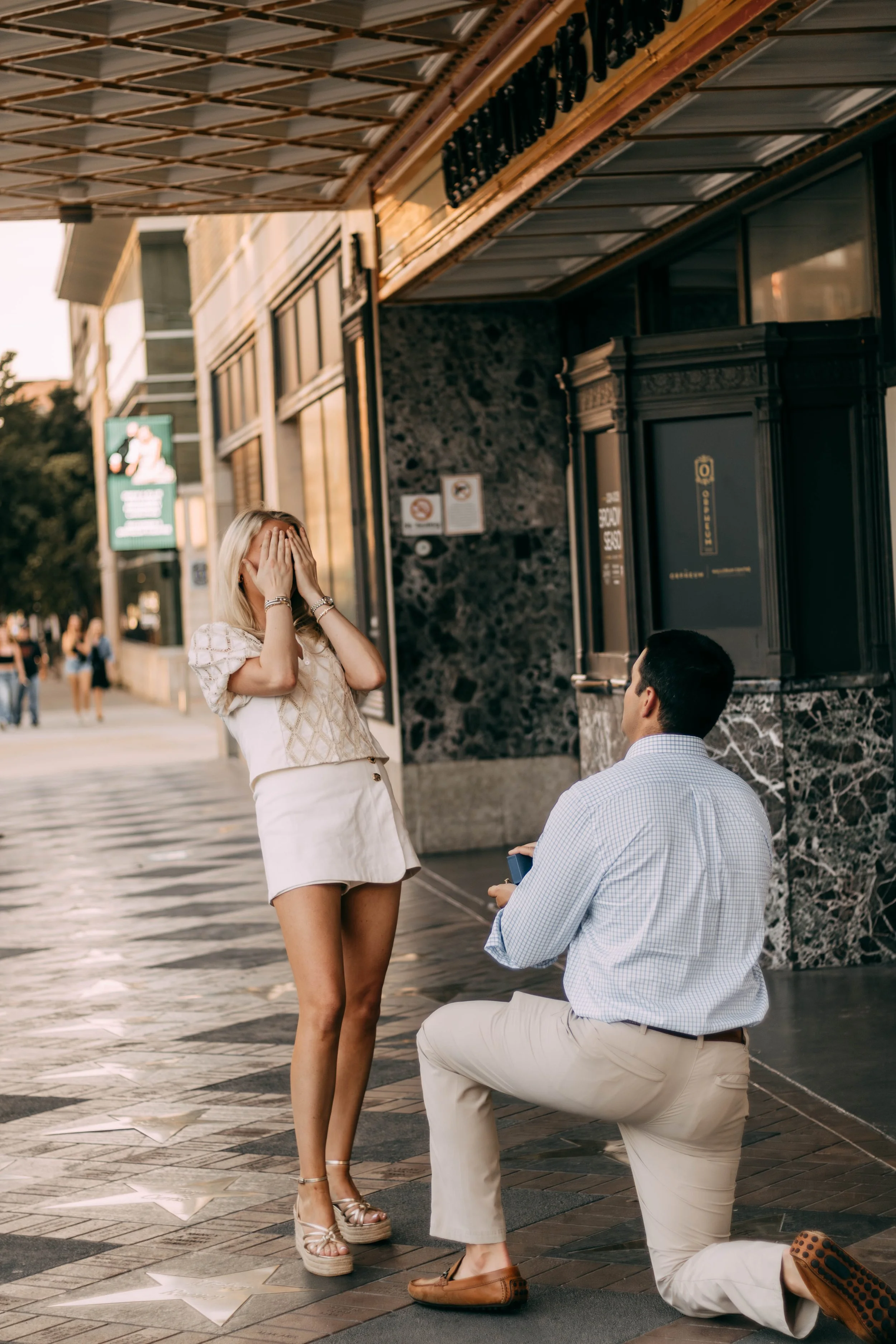 A man proposing to a woman on a city sidewalk, woman covering her face with her hands, man kneeling with a ring box, at night with city lights.