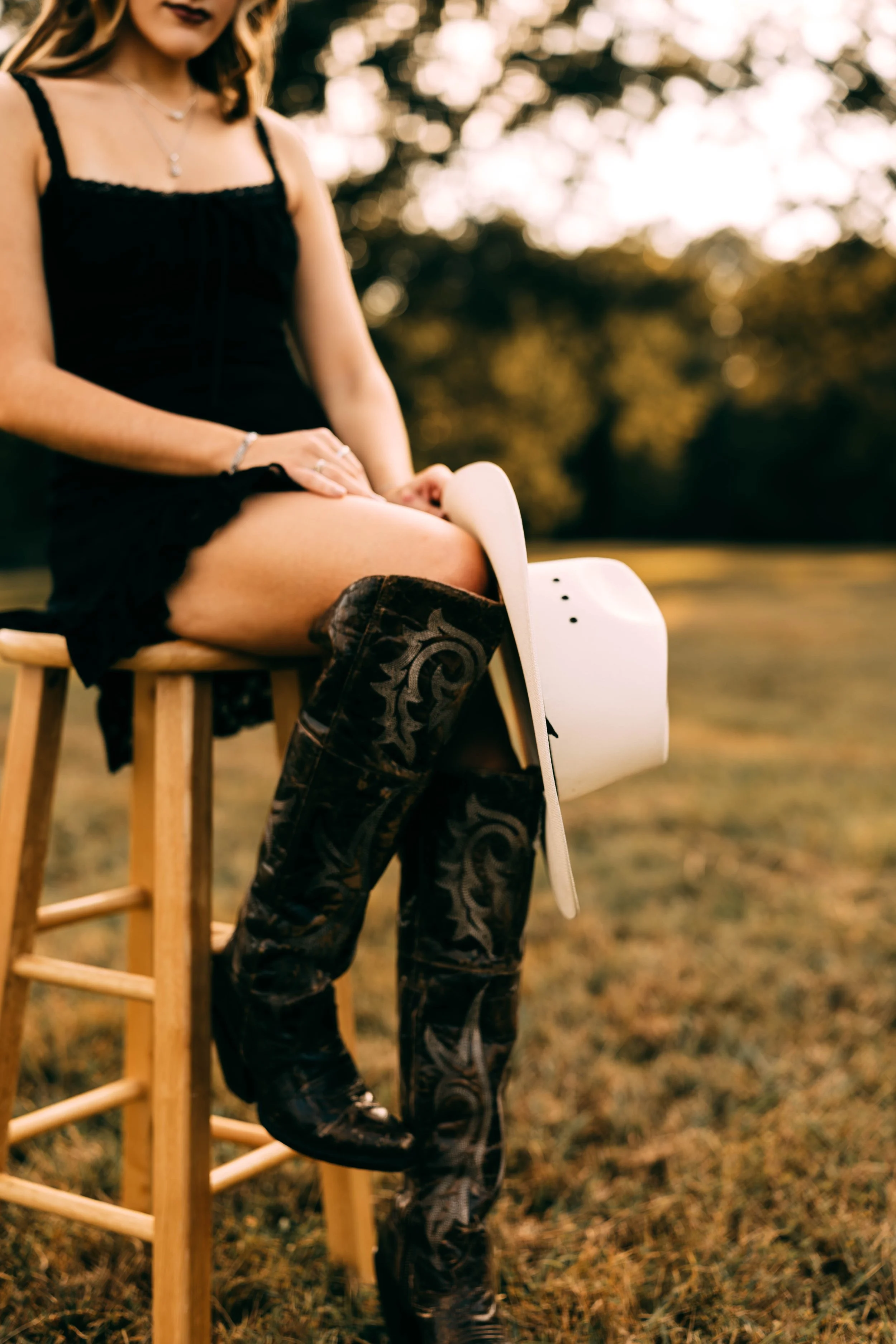 A woman sitting on a wooden stool outdoors with a helmet resting on her lap, dressed in a black dress and cowboy boots, during sunset.