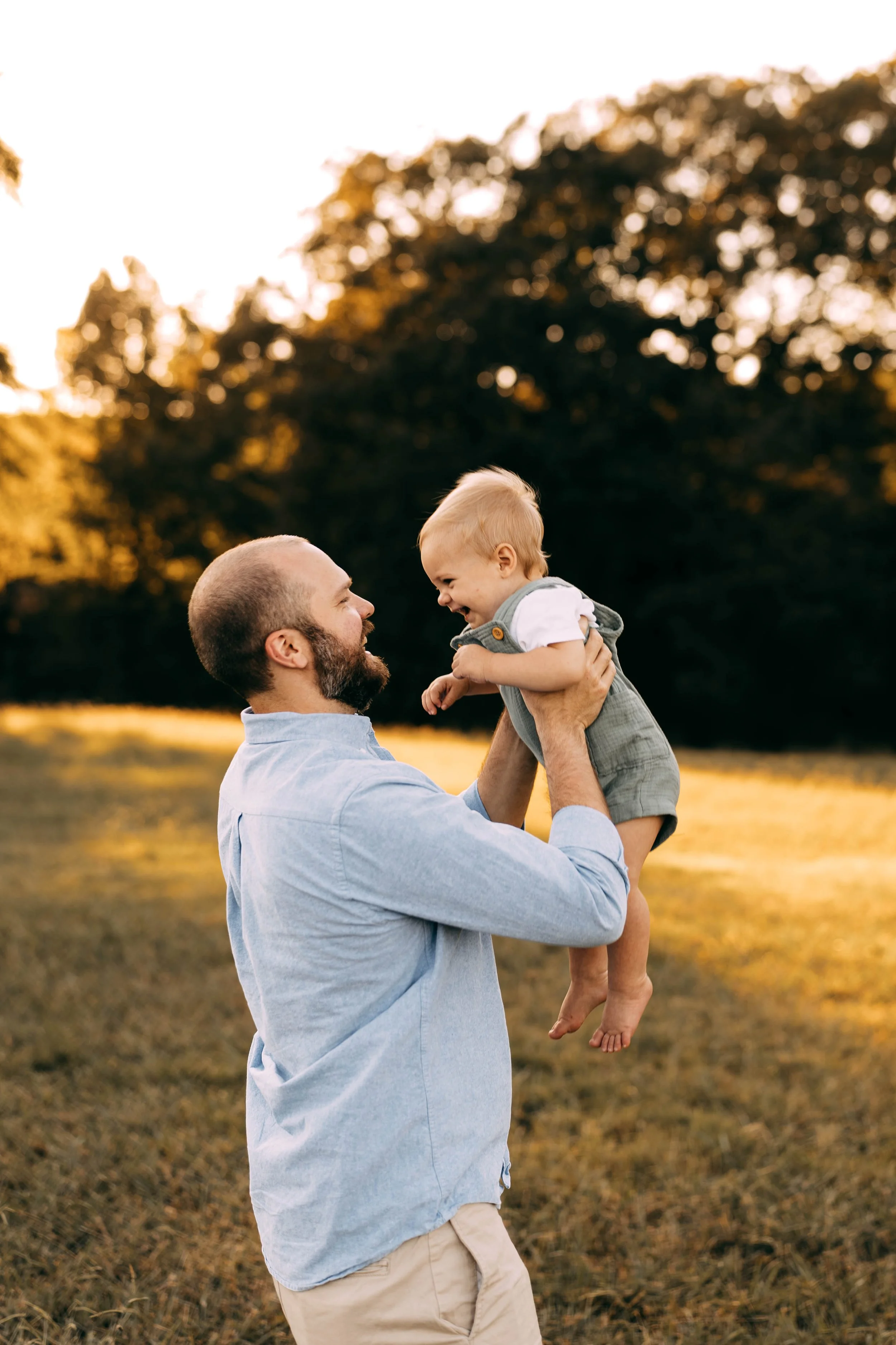 A man is holding a laughing toddler boy in a park at sunset.