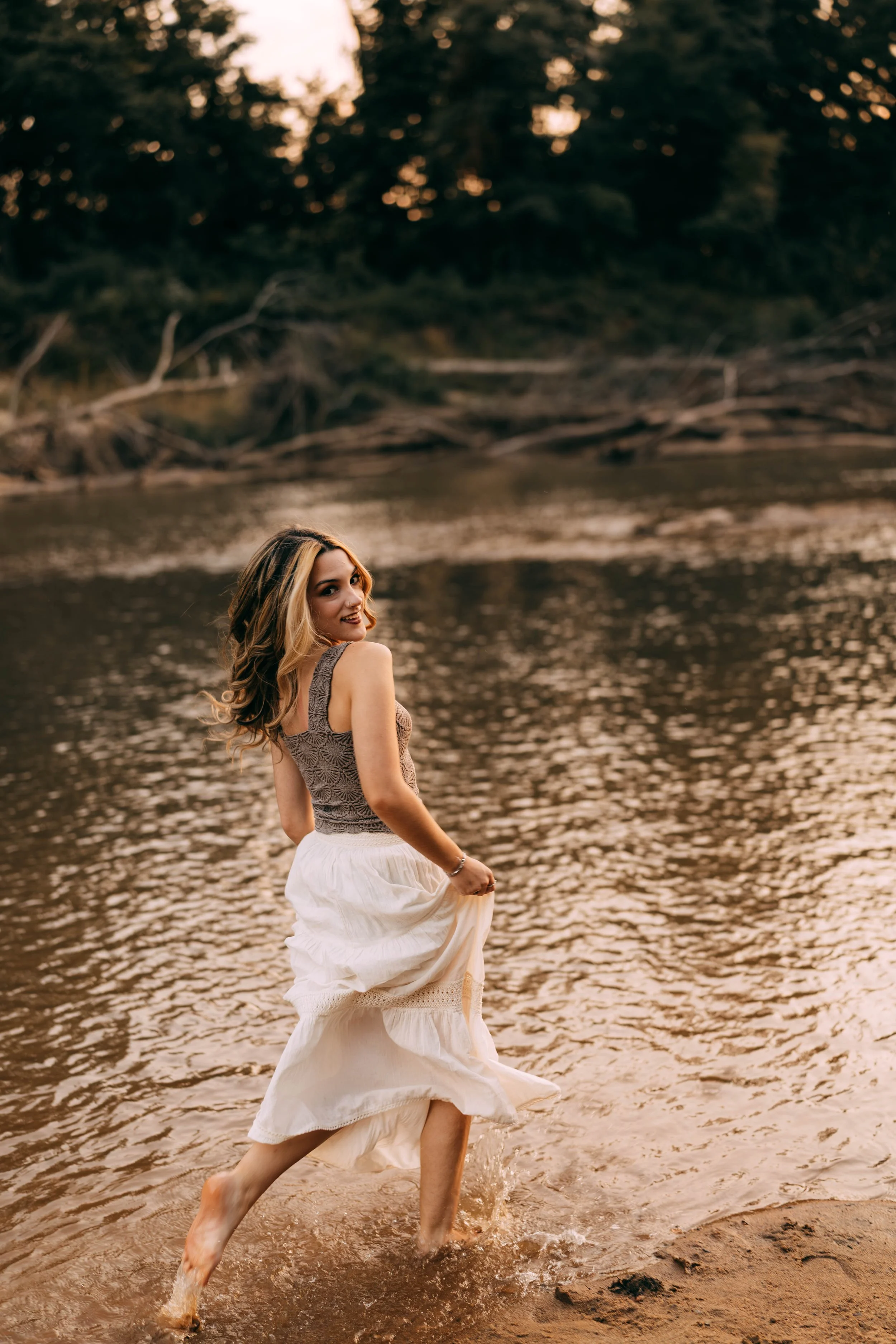 A woman in a white skirt and gray top standing in shallow water by a river at sunset, smiling and looking back over her shoulder.