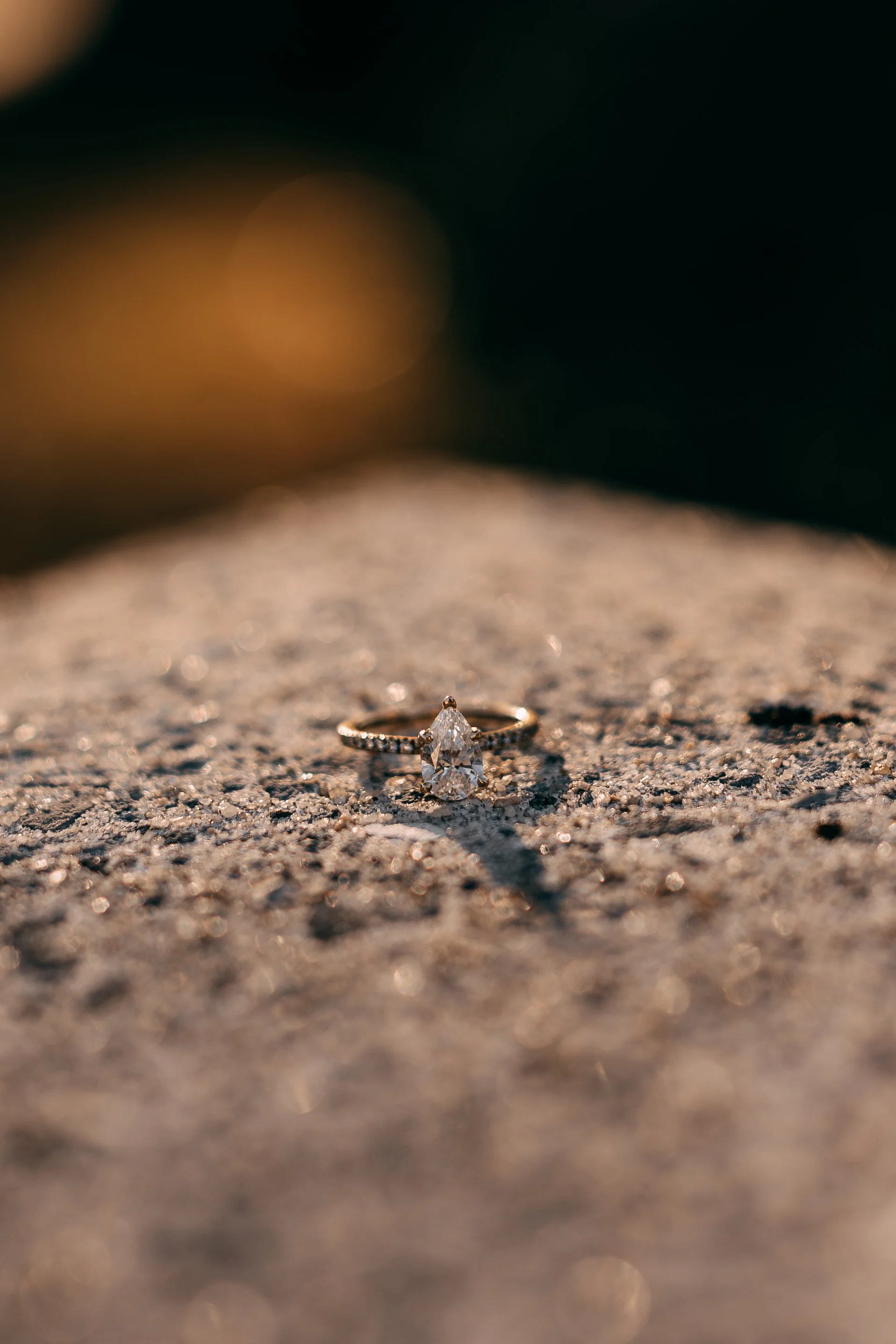 Close-up of a diamond engagement ring lying on rough, sandy surface, illuminated by warm sunlight.