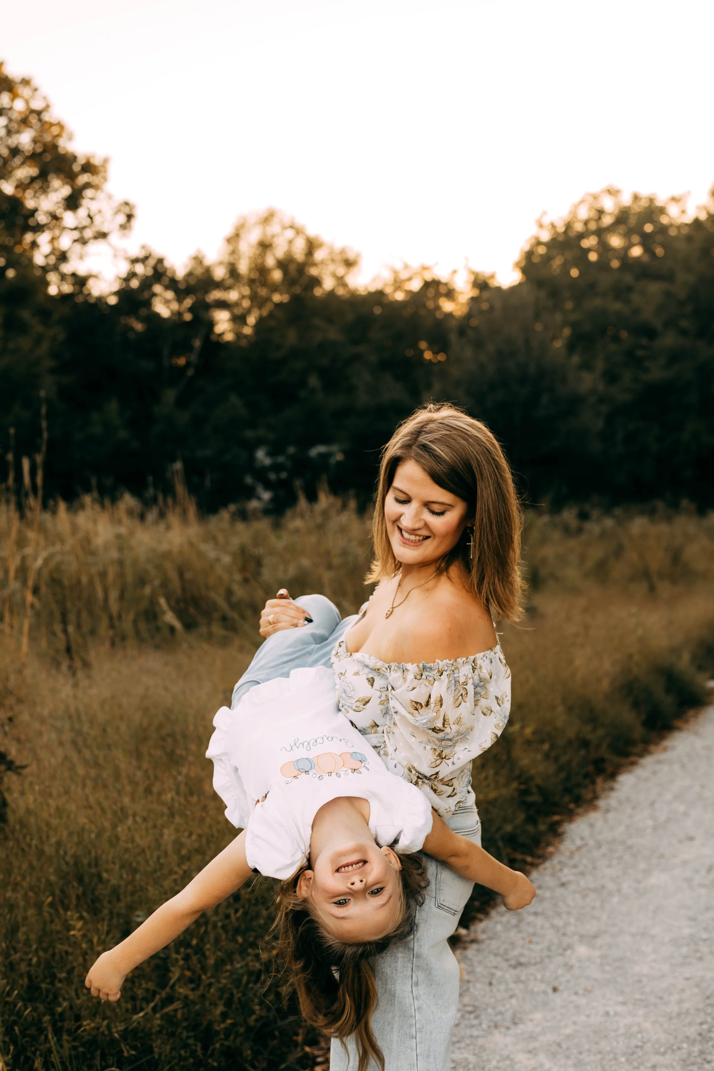 A woman holding a smiling girl upside down outdoors during sunset, with trees and a grassy path in the background.