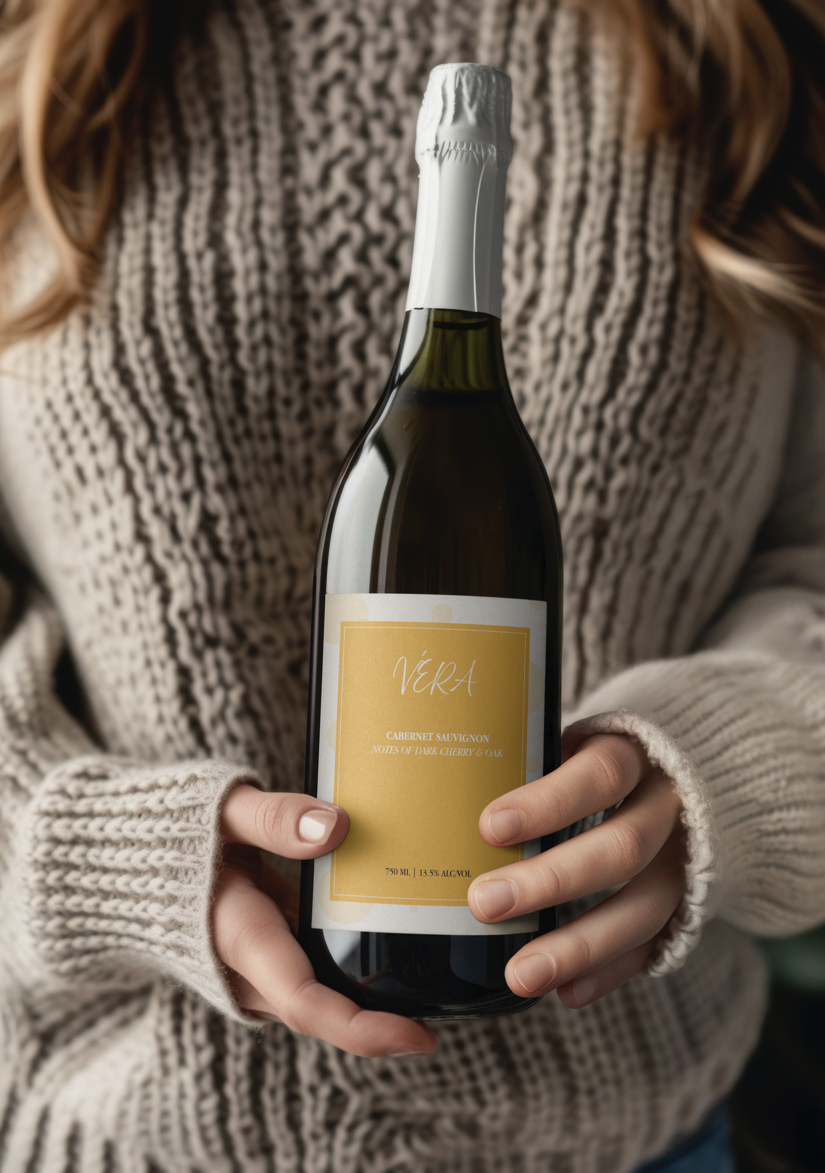 Person holding a bottle of VERA Cabernet Sauvignon wine with a yellow label and white cap, wearing a beige and brown patterned sweater.