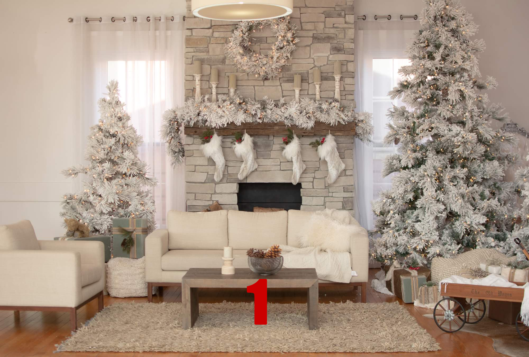Living room decorated for Christmas with two snow-covered Christmas trees, stockings hanging on a stone fireplace mantel adorned with a garland, a beige sofa and armchair, a wooden coffee table, and wrapped presents and a wagon filled with gifts