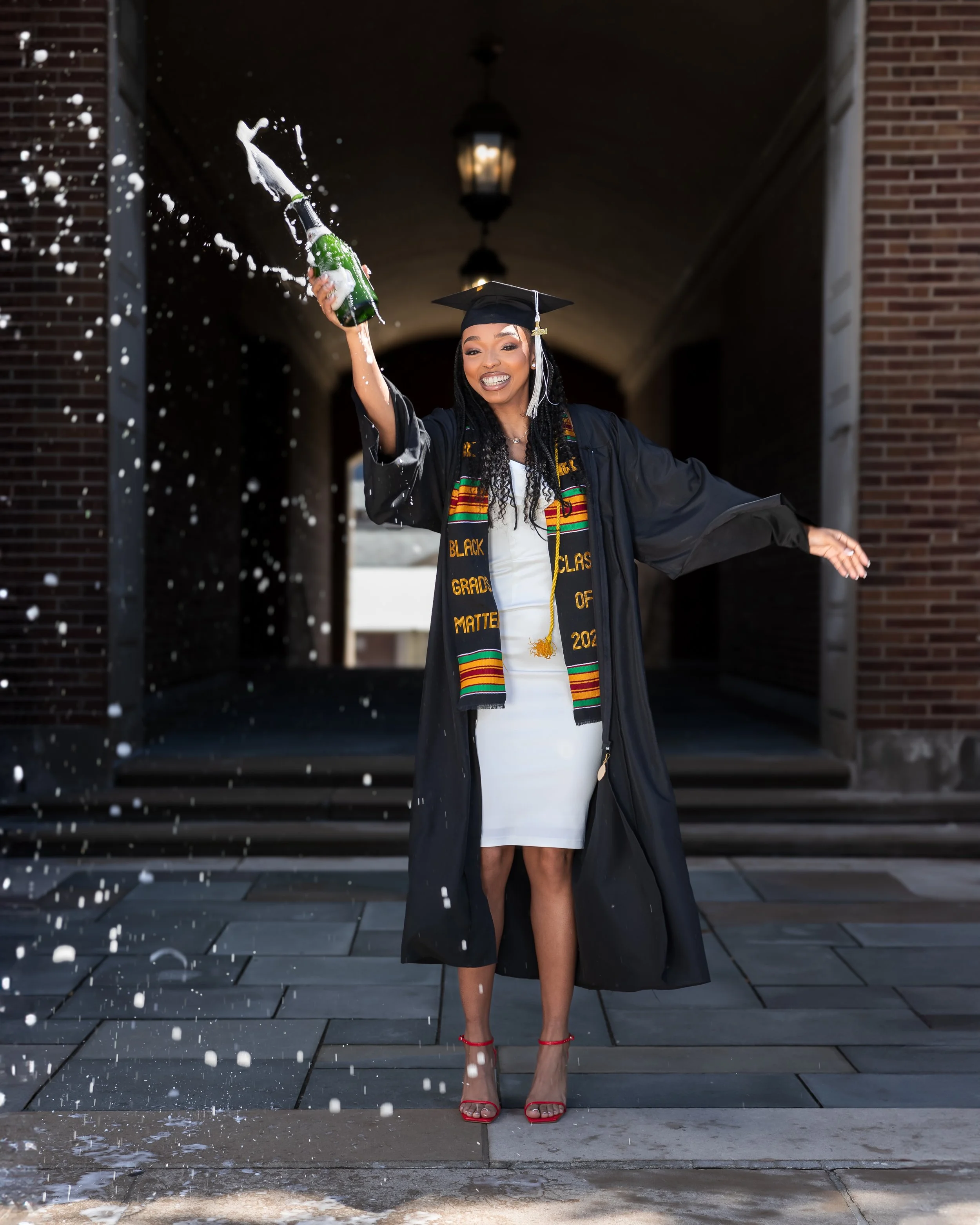 Happy woman in graduation gown and cap celebrating with champagne outside under a covered walkway.