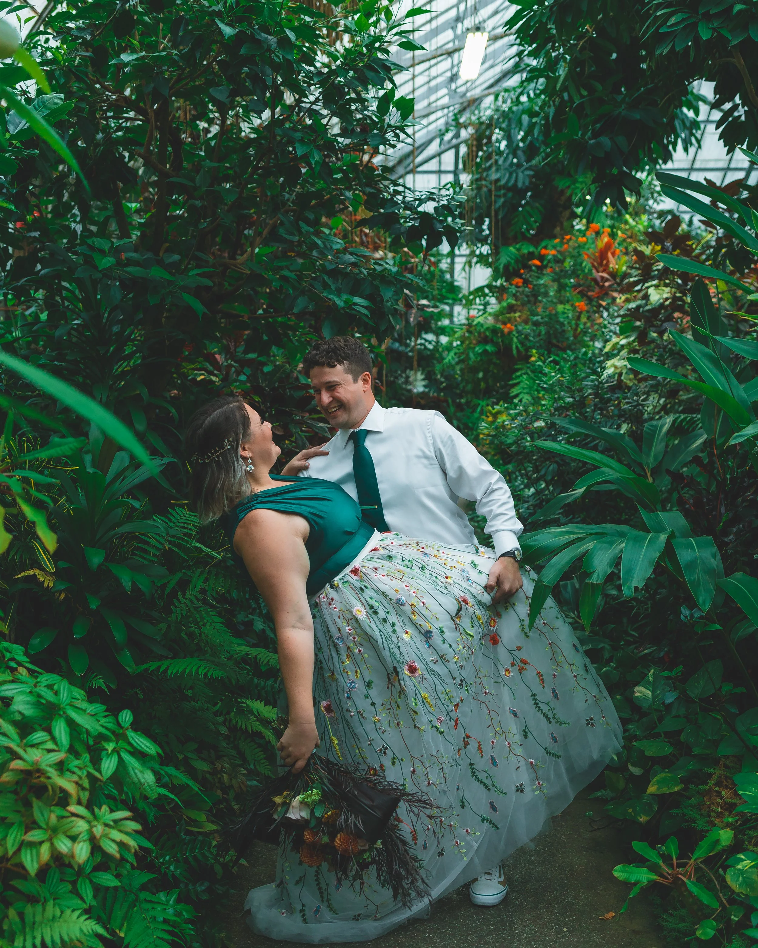 A couple in wedding attire sharing a moment in a lush greenhouse filled with tropical plants.