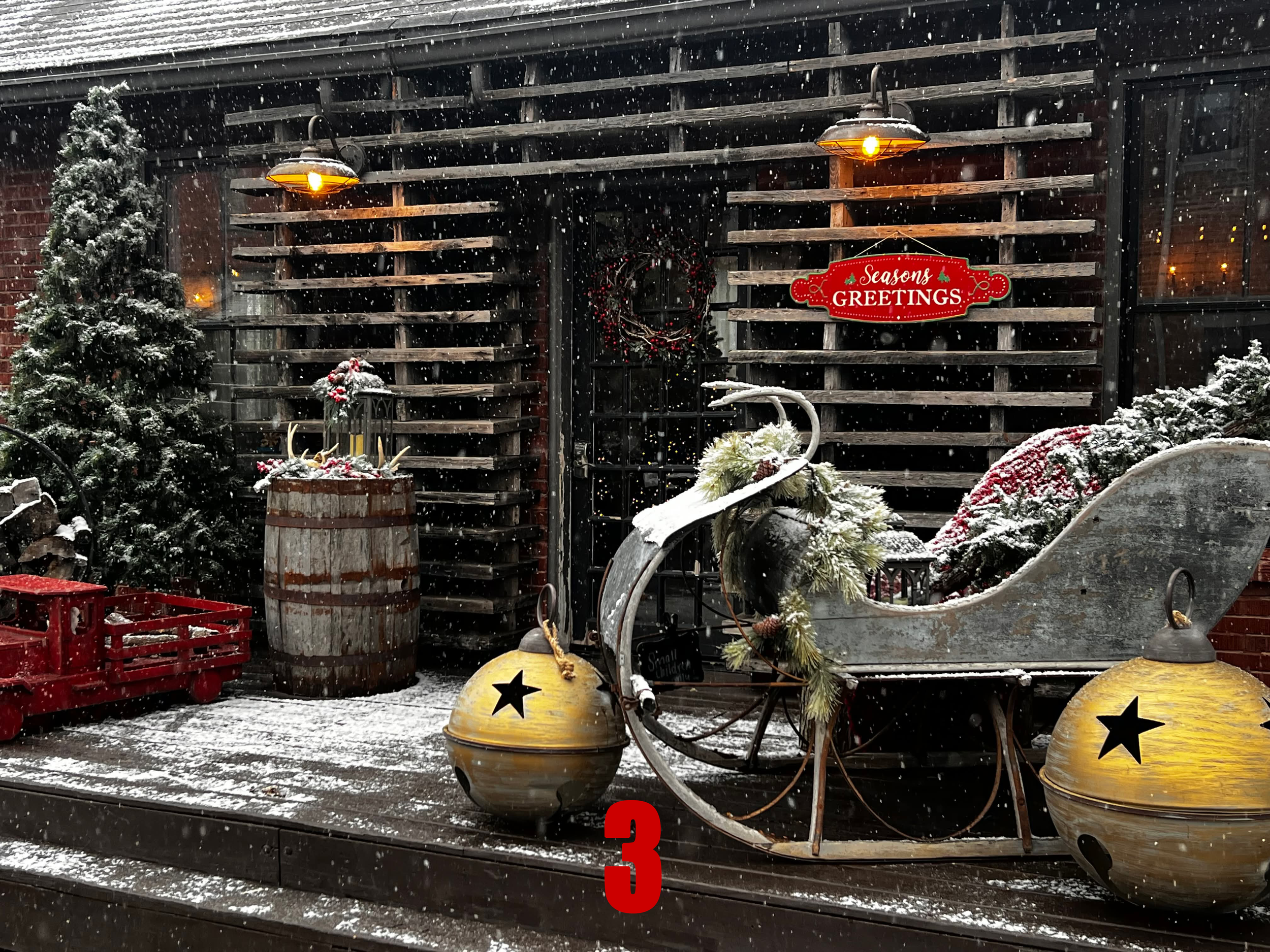 A snowy outdoor scene with a vintage sled decorated with pine branches and ornaments, a Large yellow lantern, a barrel with lanterns on top, a Christmas wreath, and a sign that reads 'Seasons Greetings,' mounted on a wooden trellis against a brick bu