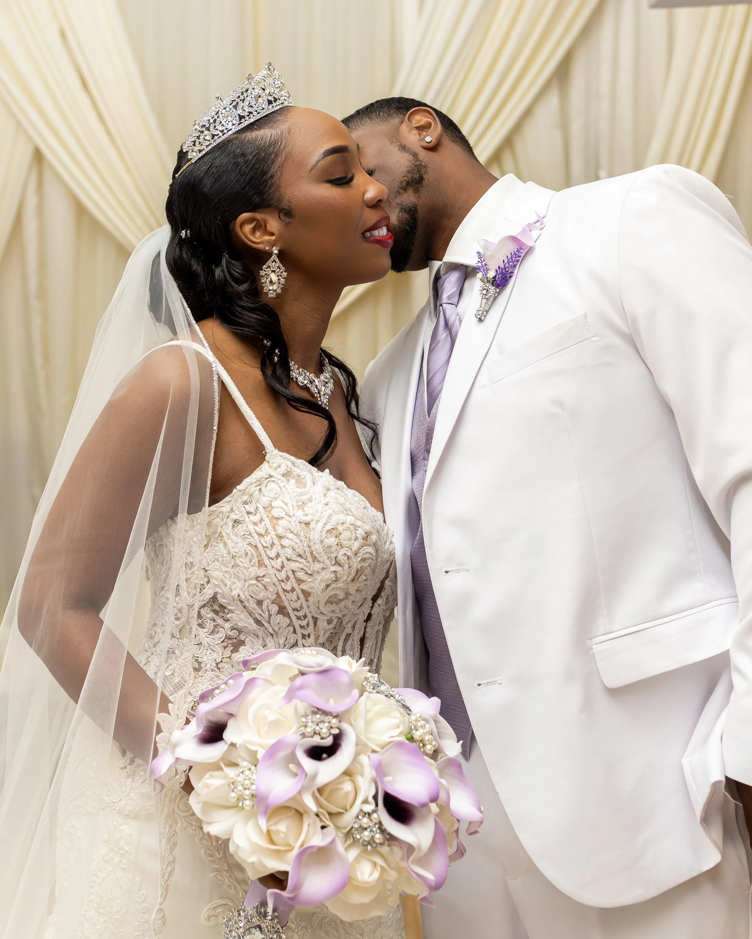 A bride and groom sharing an intimate moment at their wedding, with the bride wearing a lace wedding gown, a tiara, and holding a bouquet of white and purple flowers. The groom is dressed in a white suit with a lavender tie and boutonniere, and they 