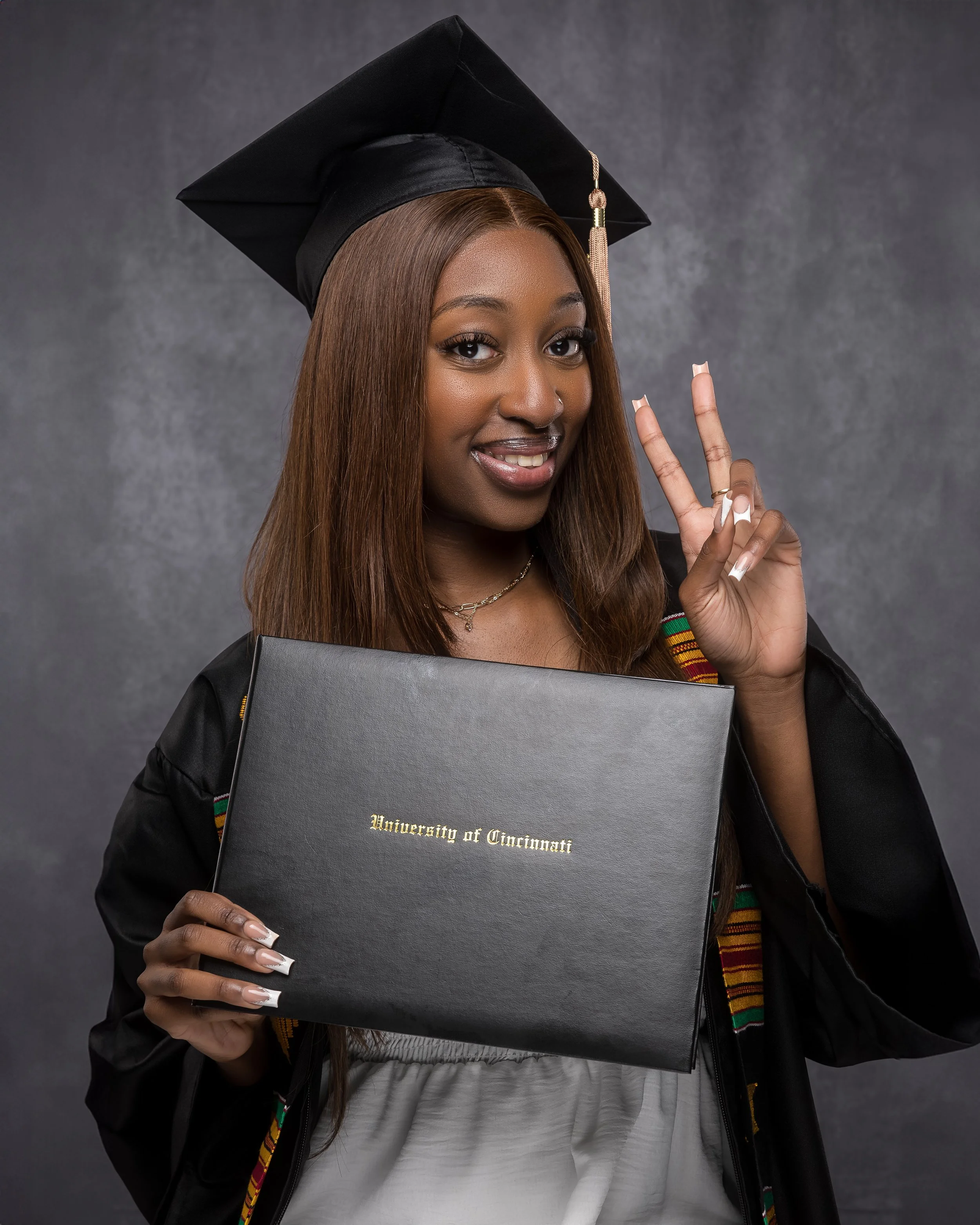 Young woman in graduation cap and gown holding diploma from University of Cincinnati, smiling and making peace sign.