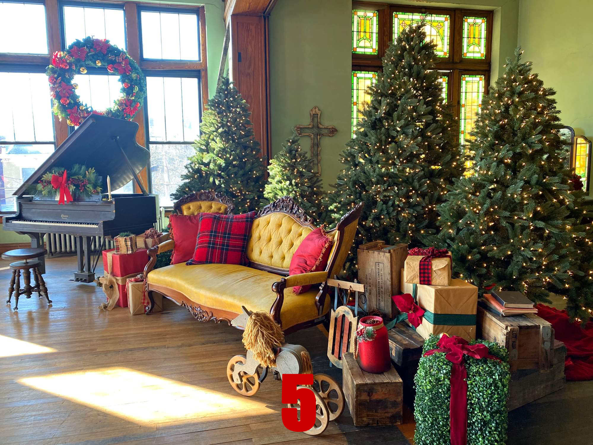 A decorated living room with three lit Christmas trees, a vintage yellow velvet settee with red and plaid pillows, a black grand piano with a Christmas wreath and red bow, various wrapped presents, a wooden rocking horse, and a large red number five 