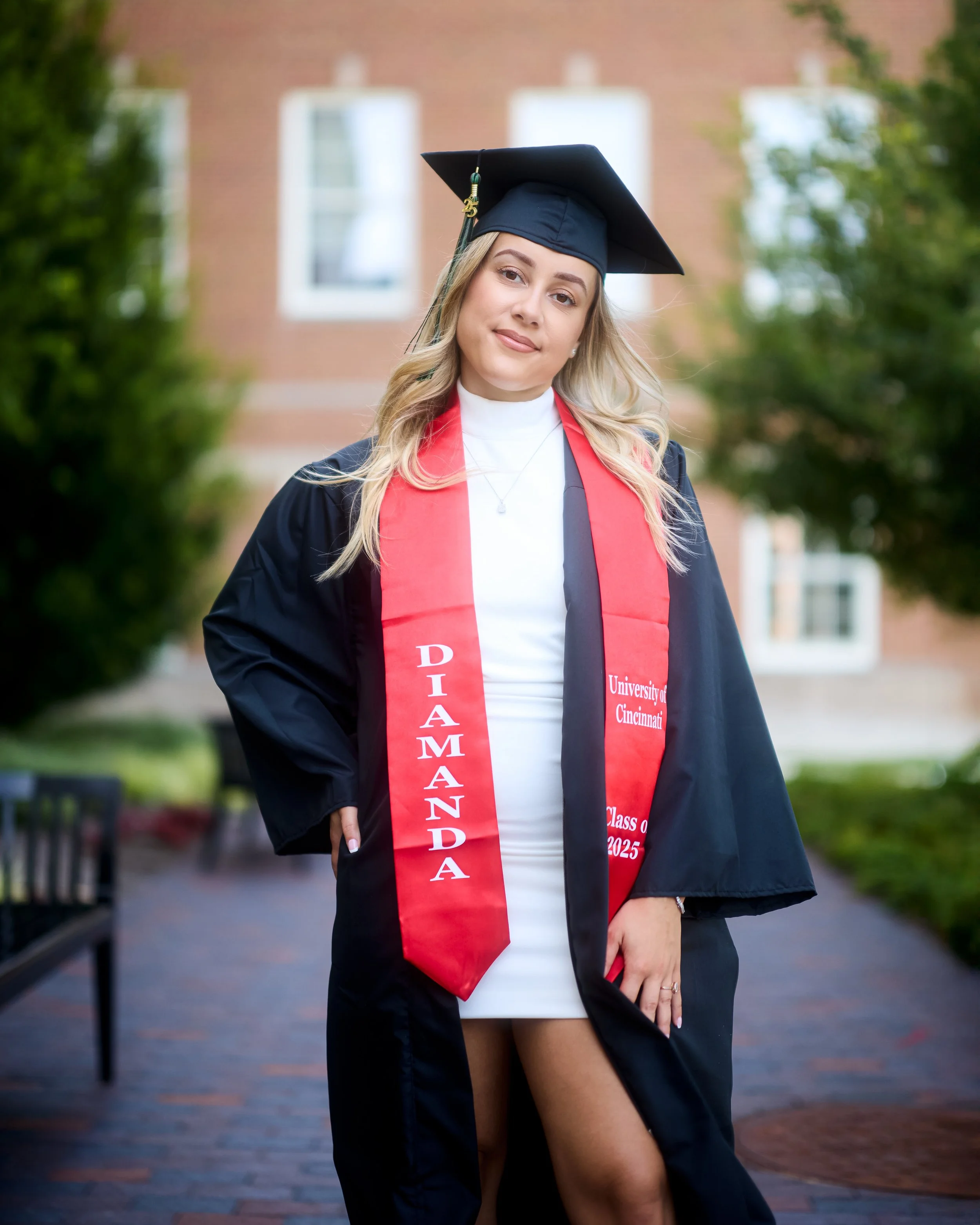 Young woman in a black graduation gown and mortarboard cap with a red stole and sash, standing outdoors on a brick pathway with greenery and a brick building in the background.