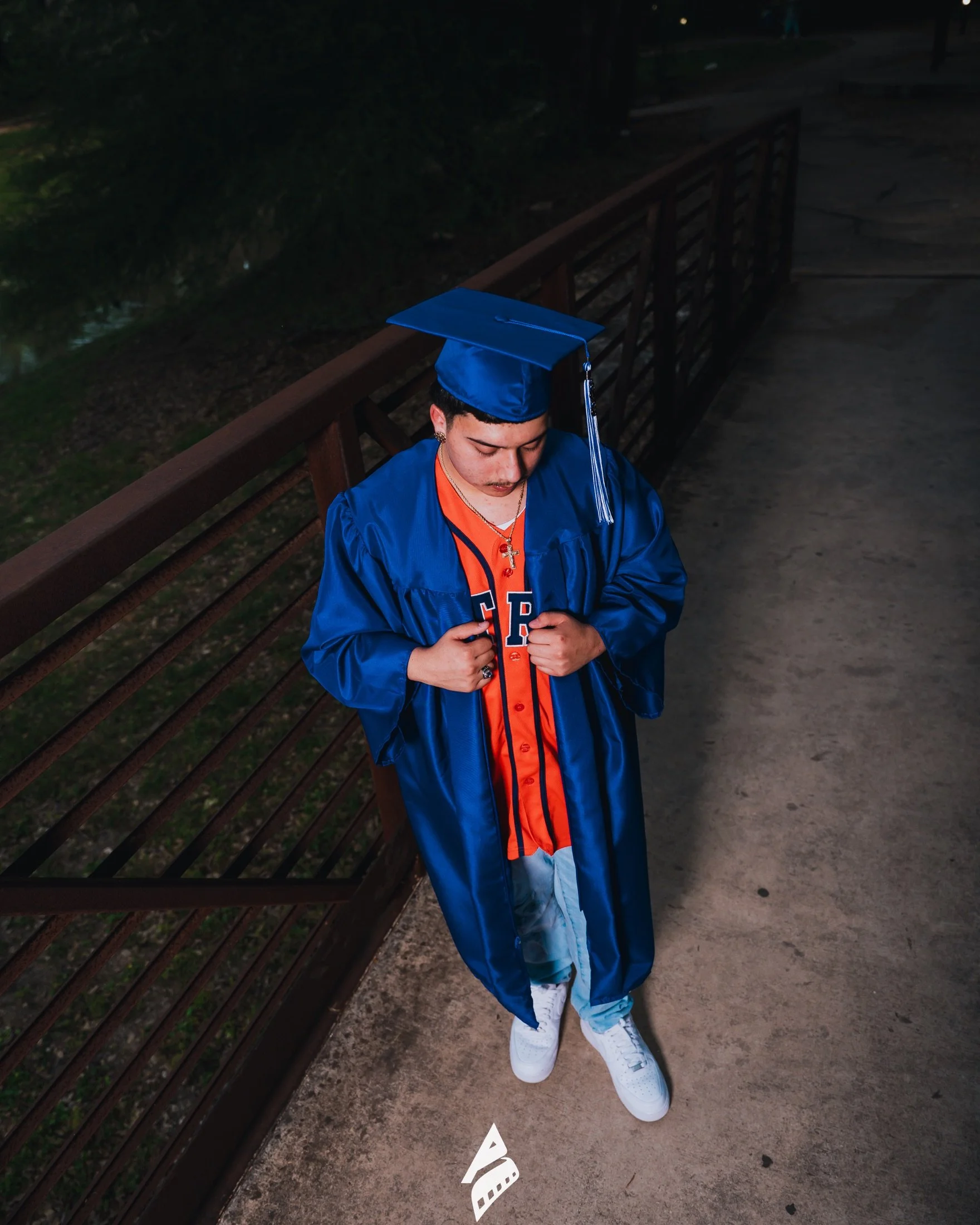 Graduate in cap and gown over an orange baseball jersey on a bridge.