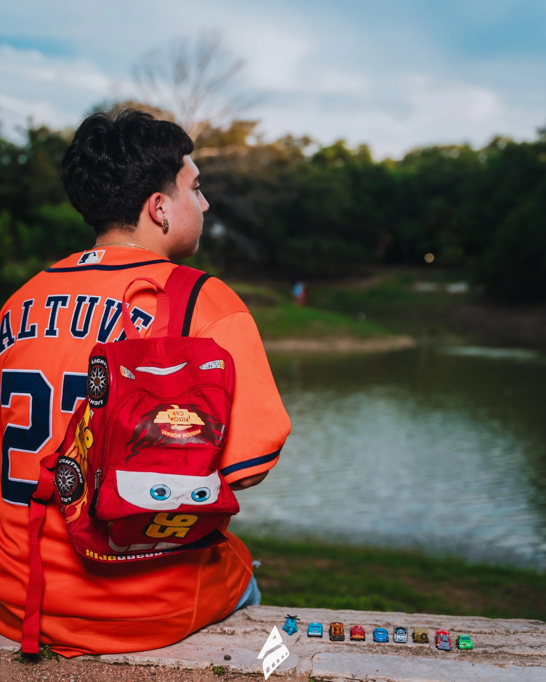 Graduate in an orange baseball jersey beside a pond, with a red backpack and toy cars.