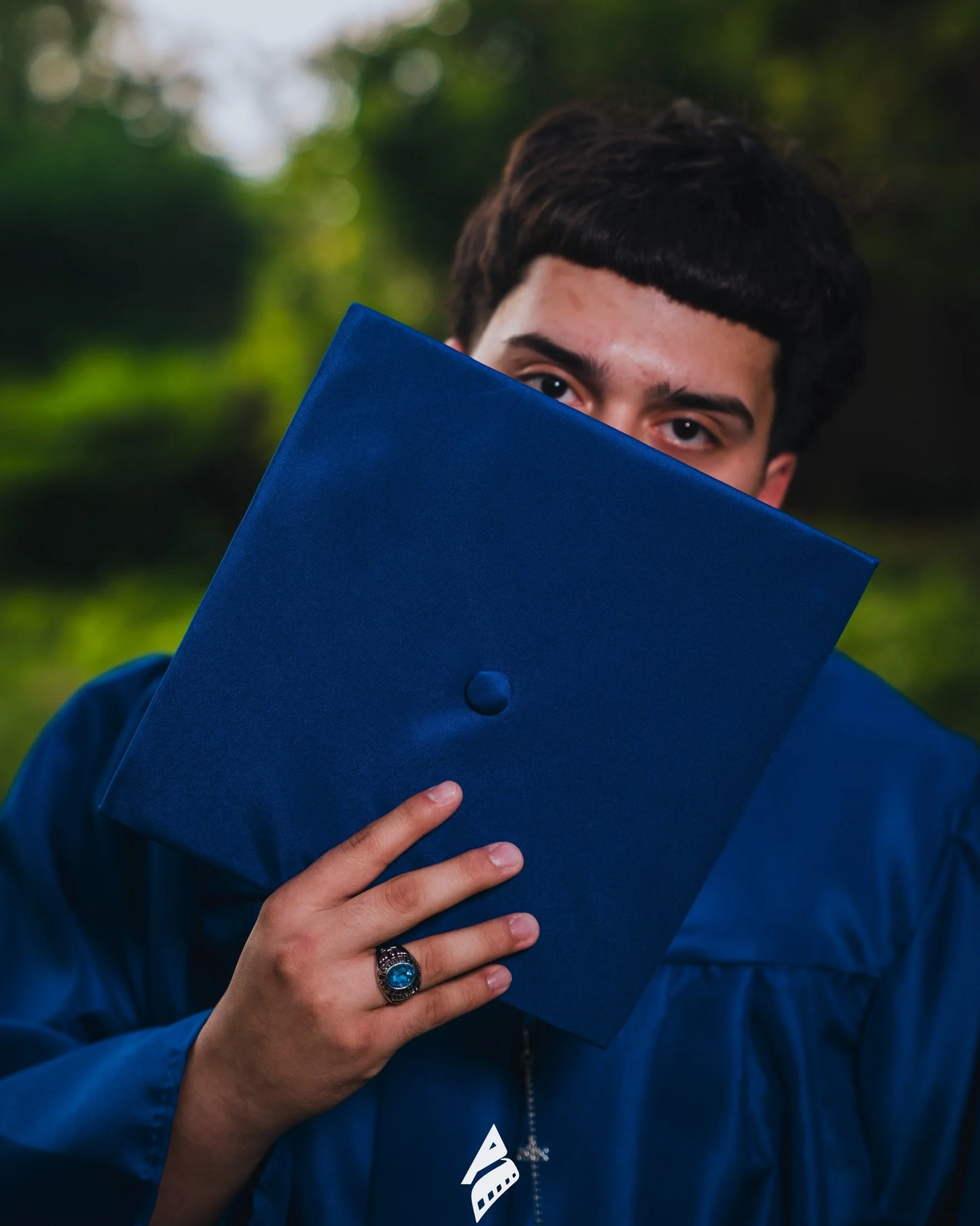 Graduate holding a blue cap in front of his face, with a class ring visible.