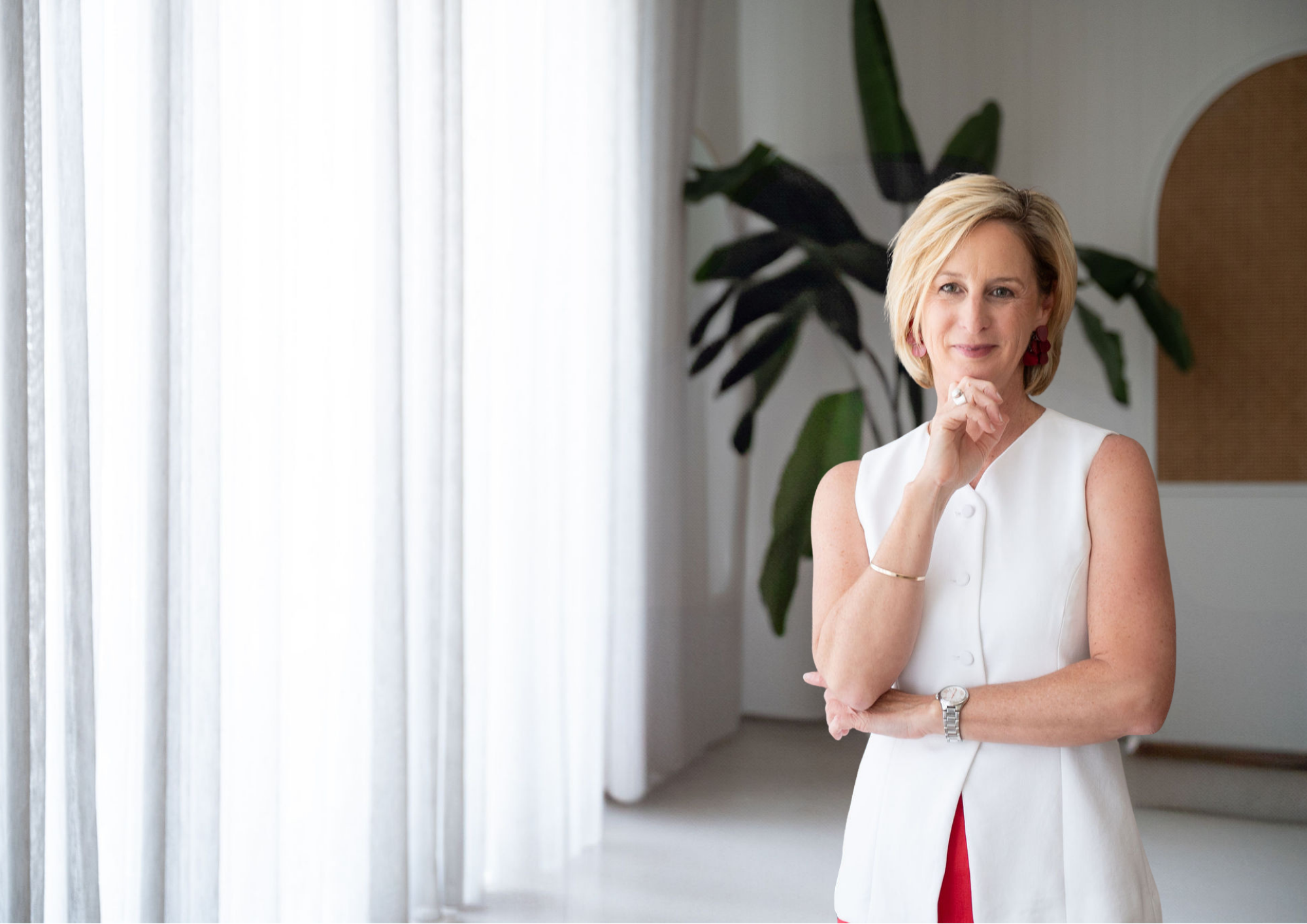 A woman with short blonde hair wearing a sleeveless white dress and red earrings standing indoors near white curtains and a large green plant.