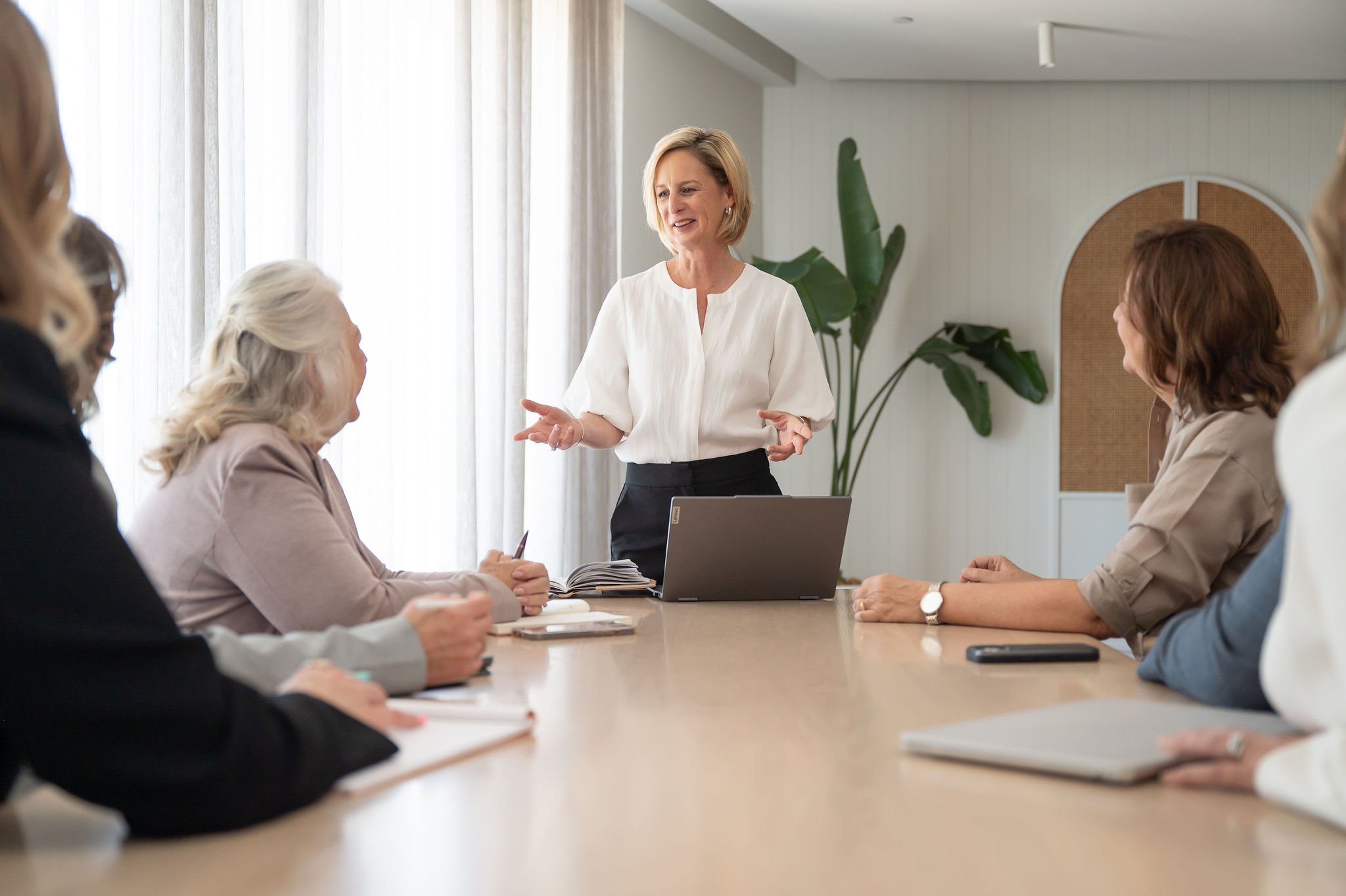 A woman giving a presentation to a group of colleagues in a conference room, with plants and white curtains in the background.