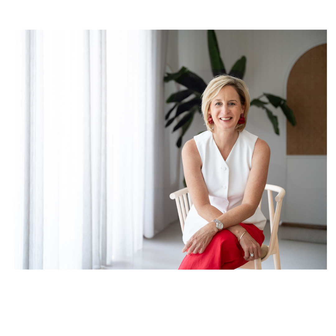 A woman with blonde hair smiling, sitting on a chair in a bright room with white curtains and green plants, wearing a sleeveless white top with red pants and earrings.