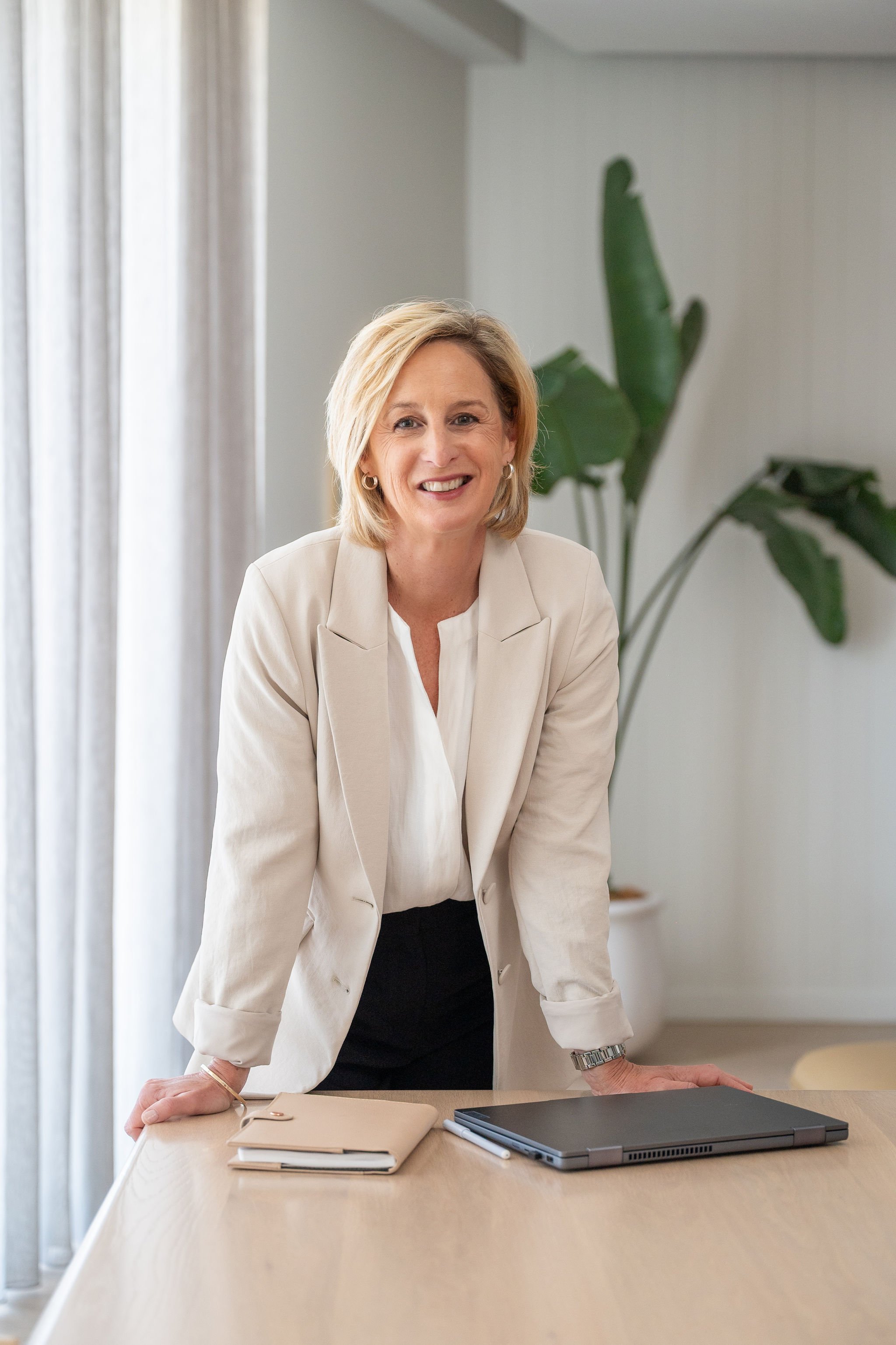 A woman wearing a cream blazer and white blouse, smiling and leaning on a wooden conference table in a modern, well-lit office with a large green plant in the background.
