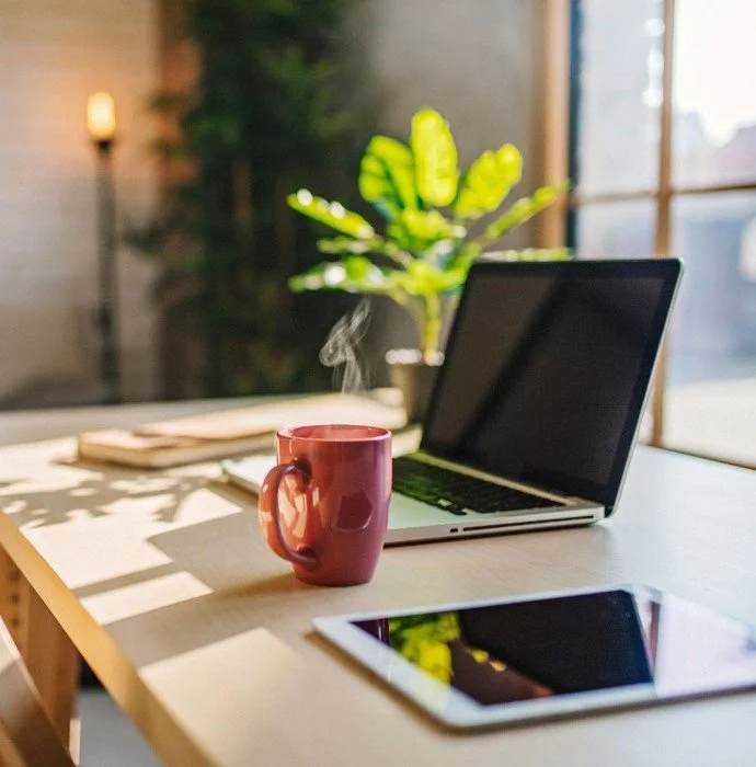 A workspace with a steaming cup of coffee, an open laptop, and a tablet on a wooden table, with a green plant and windows in the background.