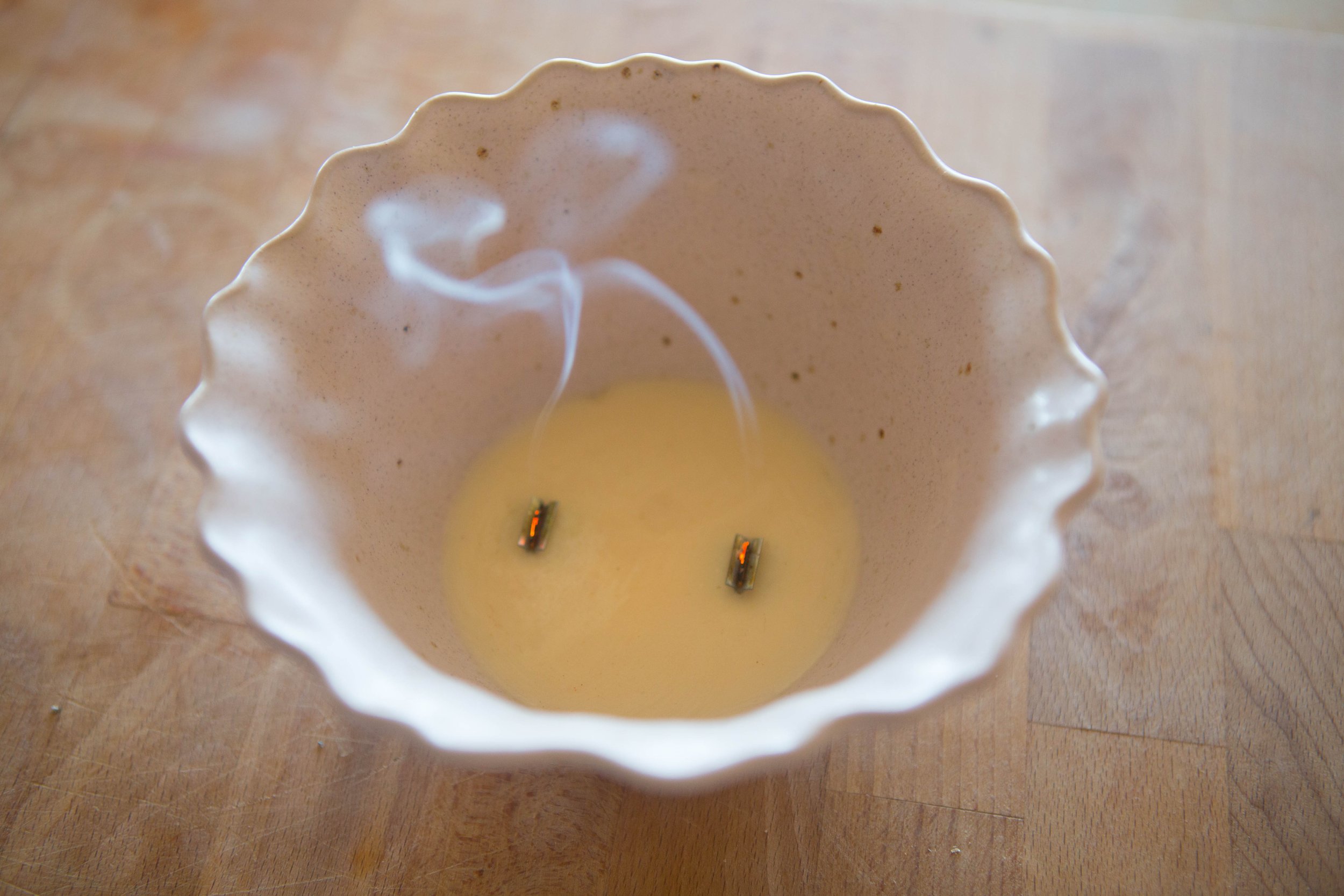 A close-up of a burning candle inside a white ceramic holder with a fluted edge, with visible smoke rising from the wick on a wooden surface.
