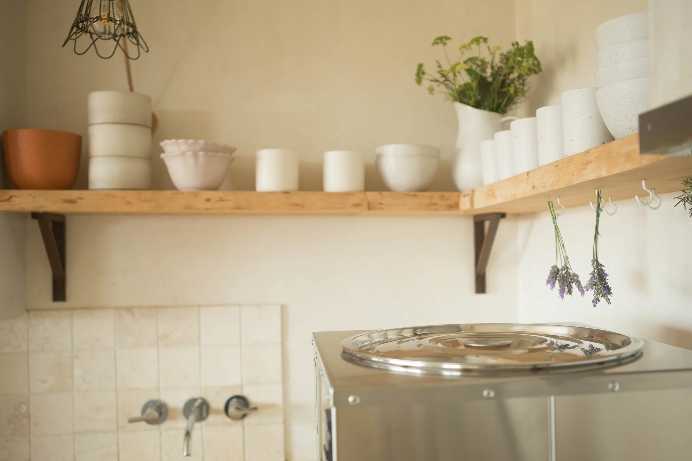 Kitchen shelf with white and terracotta bowls, cups, and a white vase with lavender flowers, above a stove with two lavender sprigs hanging, wall with tiled backsplash and control knobs.