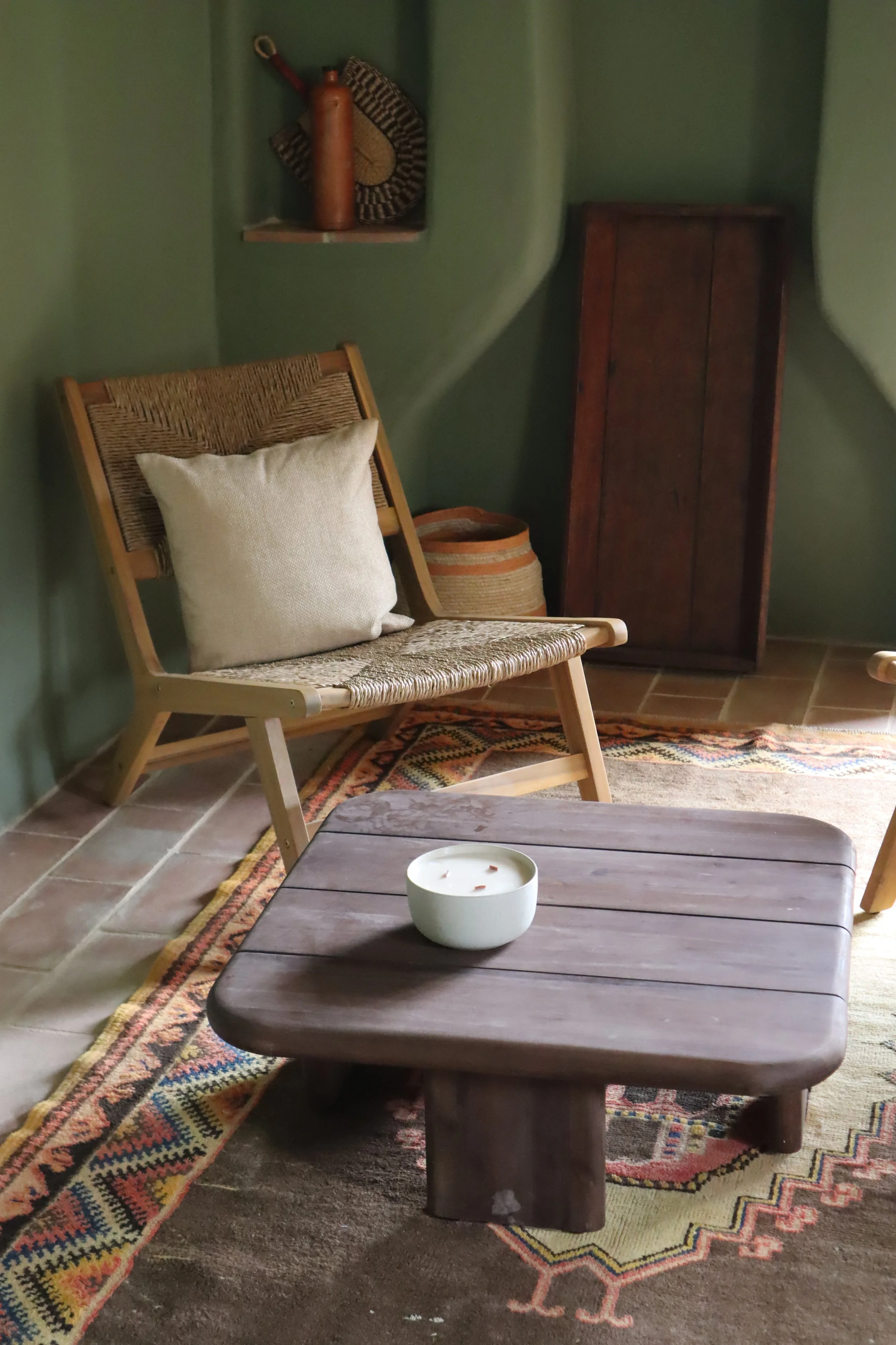 A cozy living room corner with a wooden chair with a woven seat and a cream pillow, a low wooden coffee table with a white bowl ceramic with a candle, a colorful rug, a wooden cabinet, a shelf with a carved bottle and a woven fan, and a green wall.