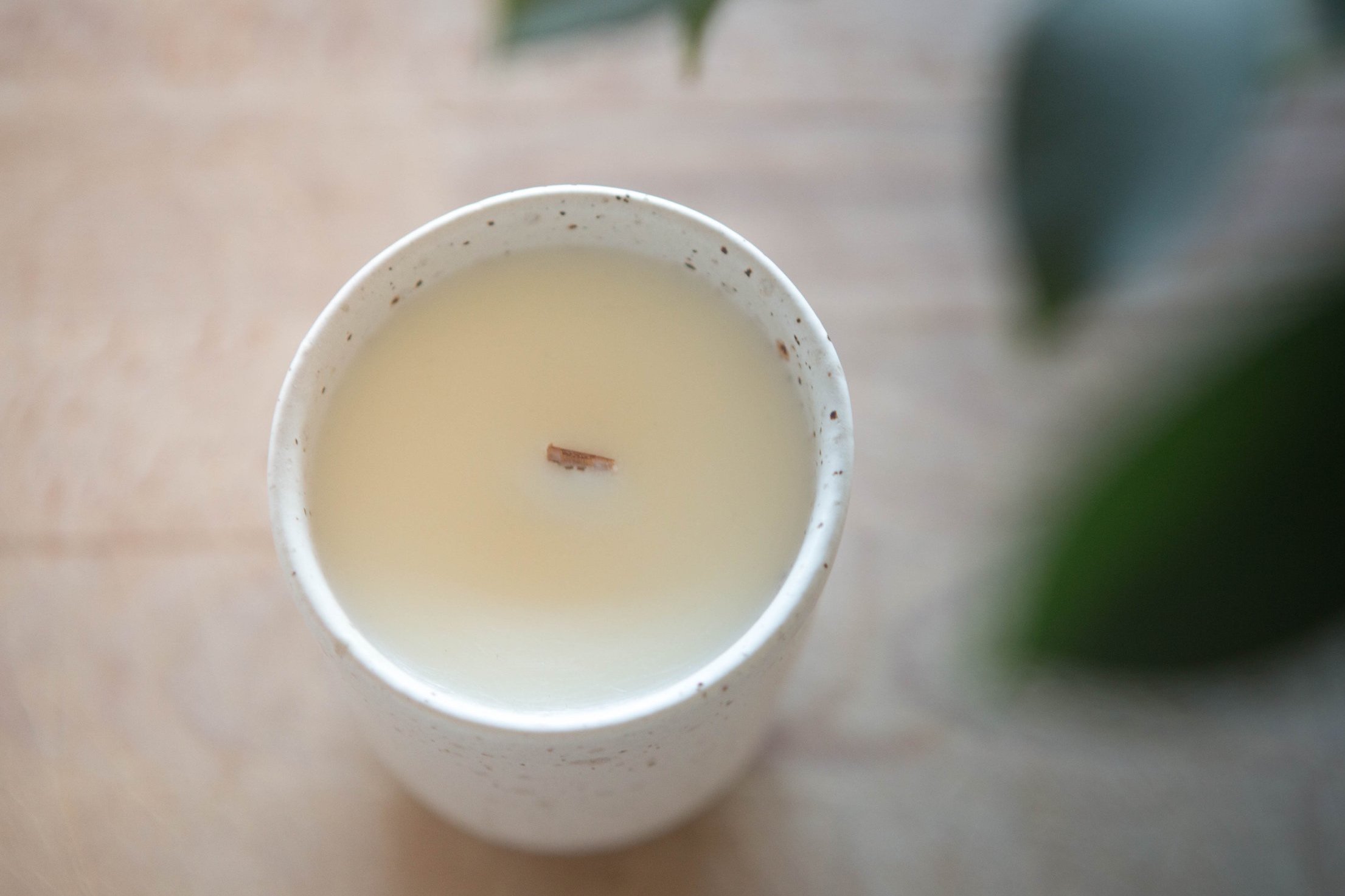 A white ceramic candle holder with a lit candle, a small wooden wick, placed on a wooden surface, partially obscured by a green plant leaf on the right.