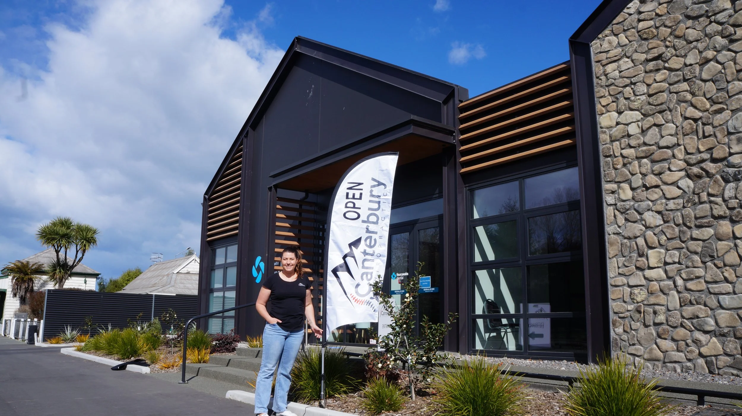 Woman standing outside the entrance of a modern building with a banner that reads 'Open Canterbury.' The building has a stone wall and large glass windows against a blue sky with some clouds.