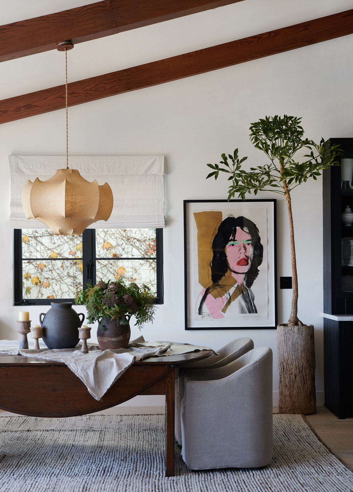 Warm modern dining room with vintage wood table, pop art portrait, sculptural pendant, and exposed beams.