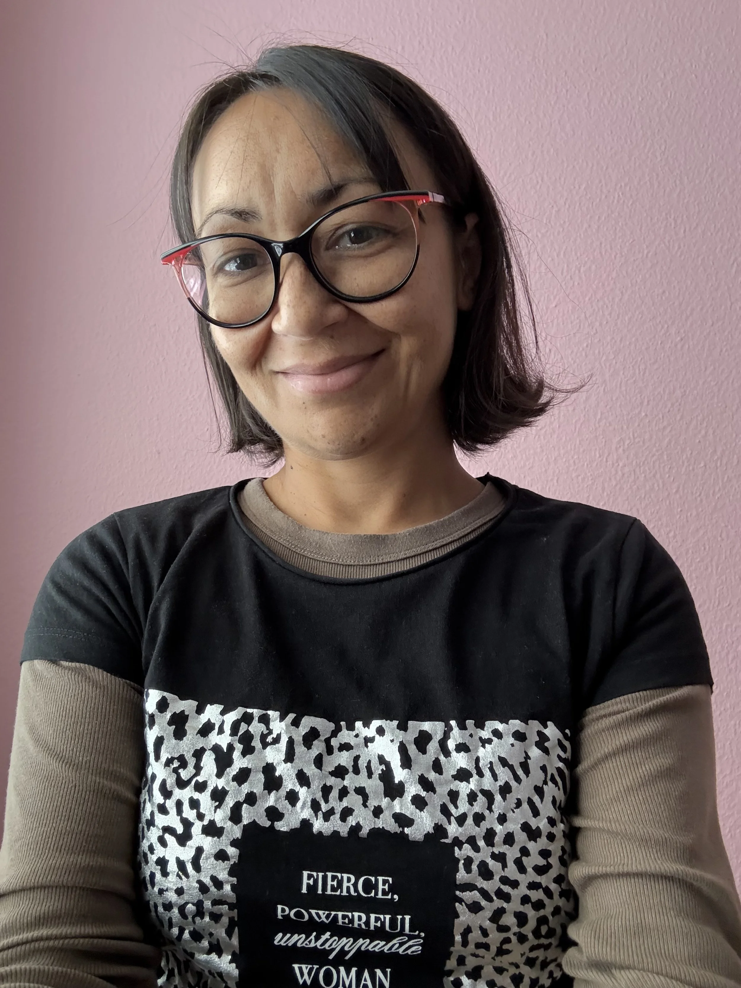 A woman with short dark hair wearing black glasses with red accents, smiling, in front of a pink wall, wearing a black t-shirt with leopard print and text that reads 'FIERCE, POWERFUL, unstoppable WOMAN.'