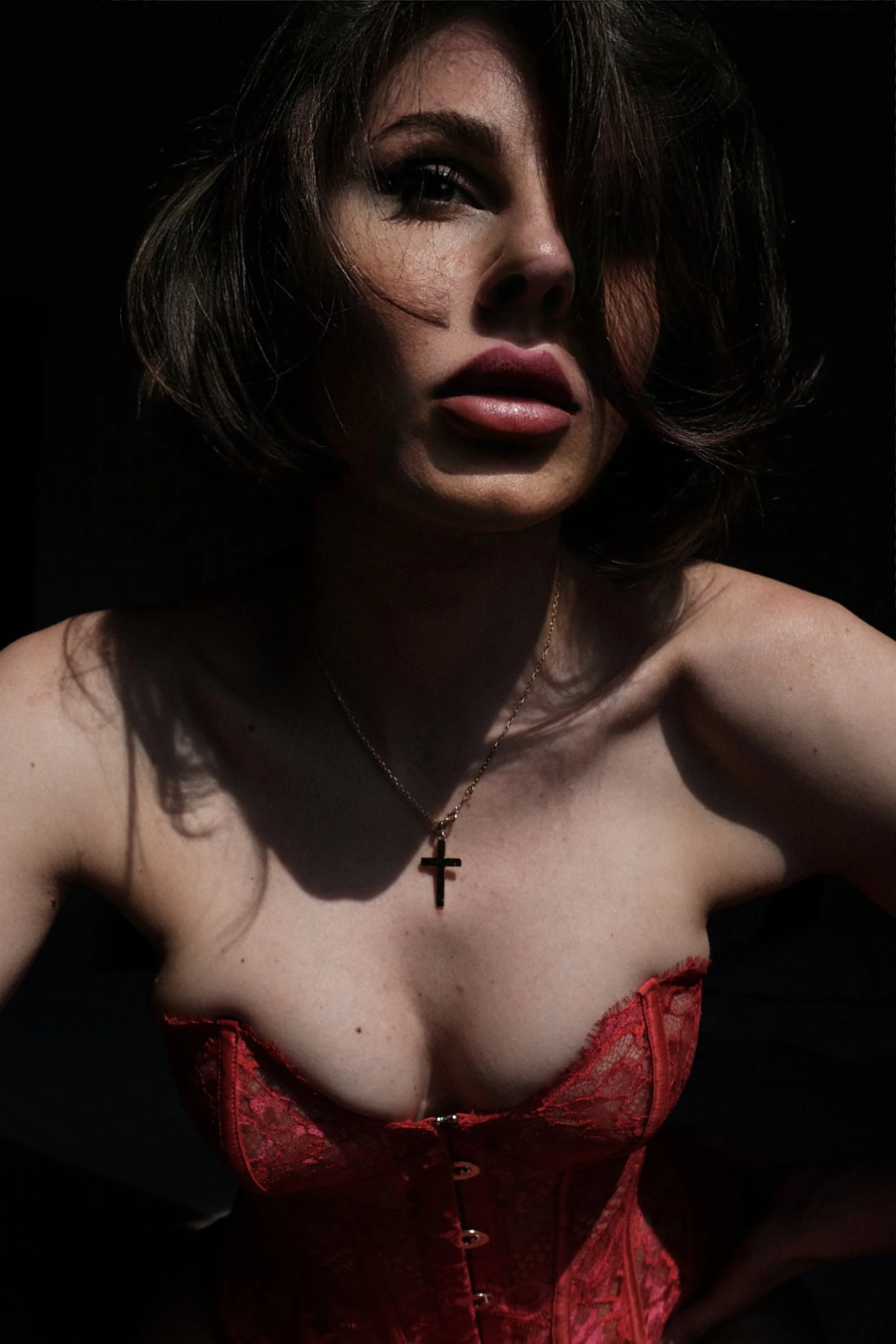 A woman with short brown hair and makeup, wearing a red lace corset and a gold cross necklace, posing against a black background.