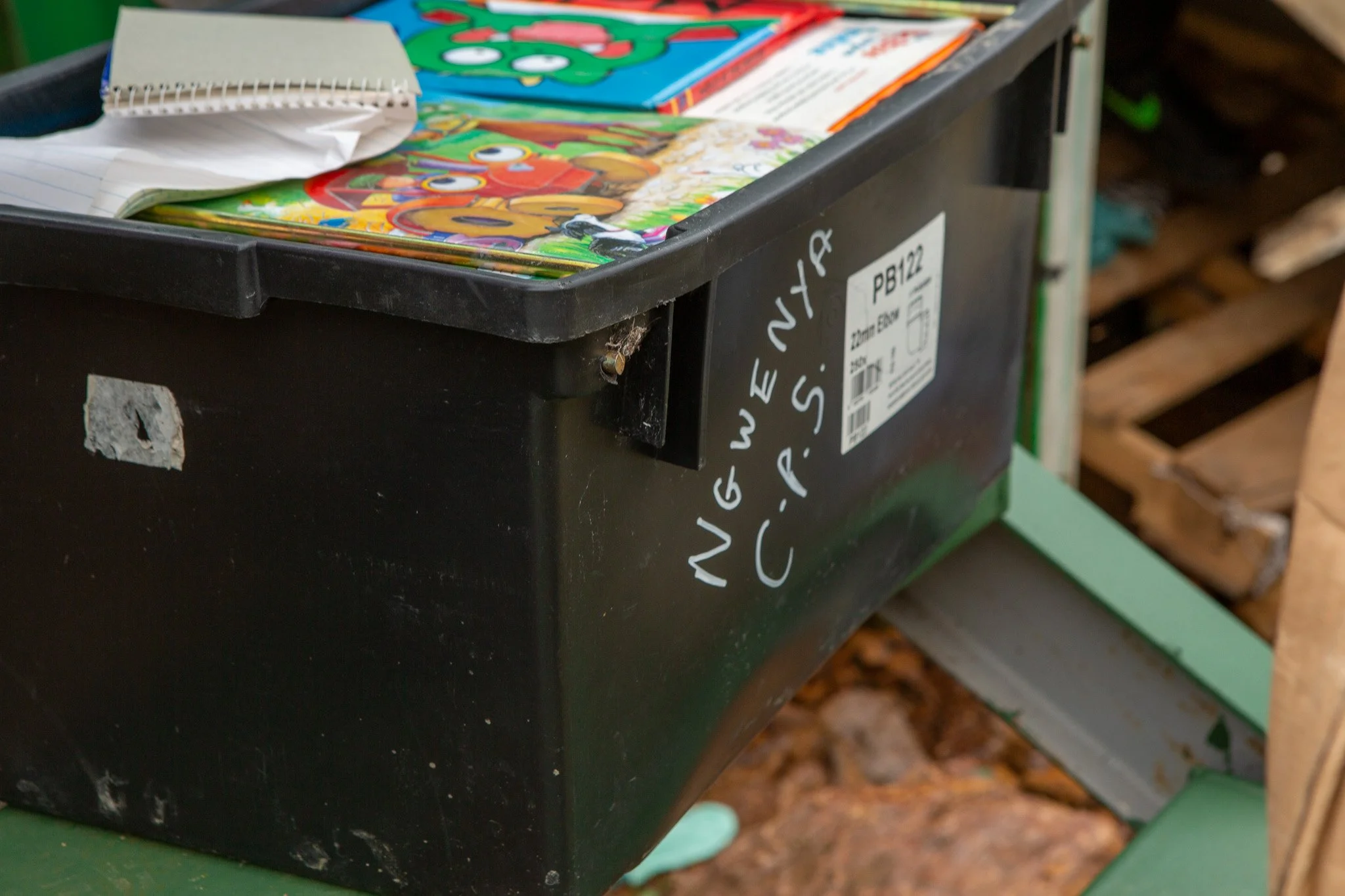 Black plastic bin labeled "Ngwenya C.P.S." containing colorful books and a spiral notebook, positioned on a green metal surface with wood pallets in the background.