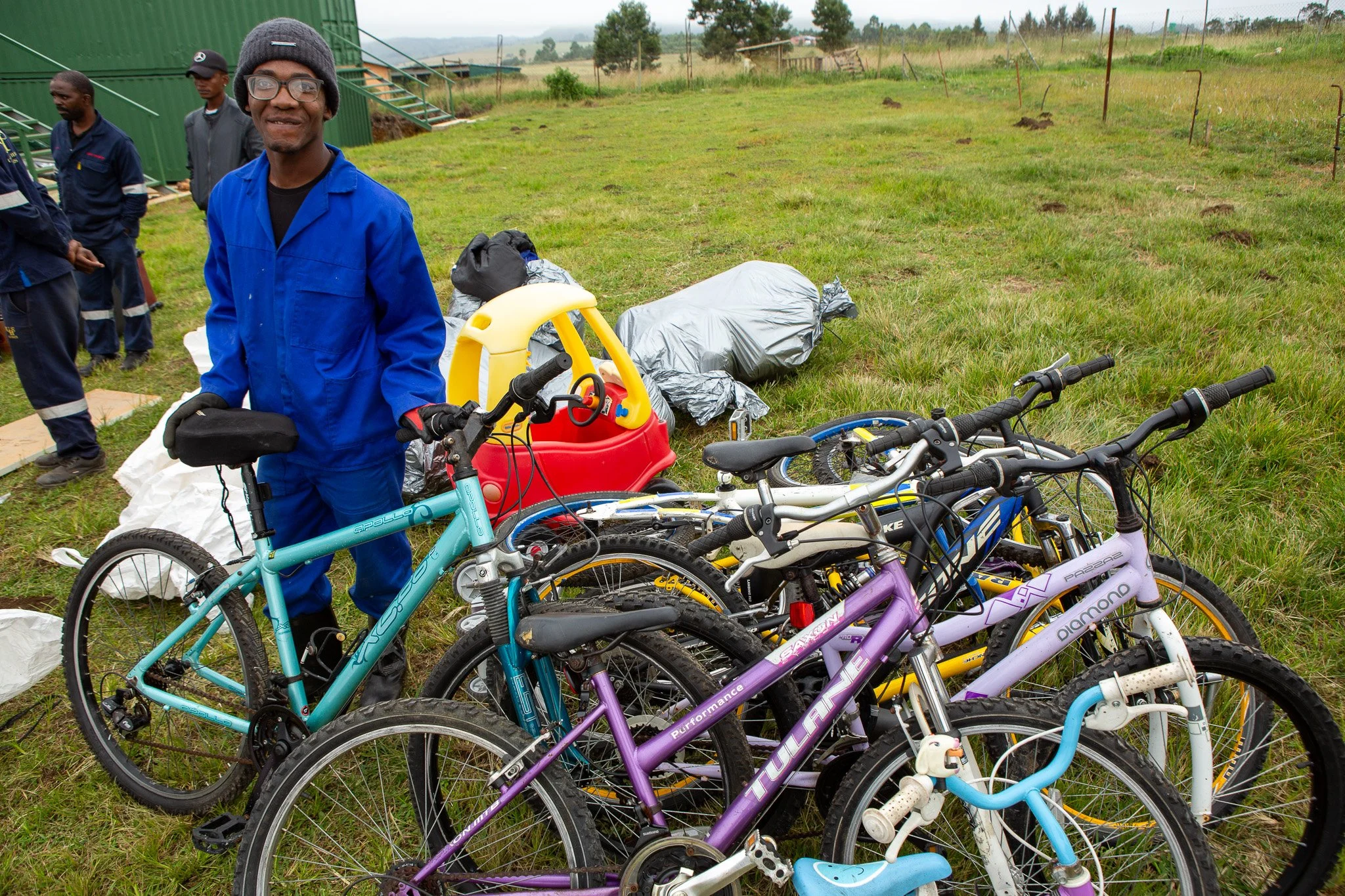 A person in blue overalls stands outside holding a bicycle among a group of bicycles on a grassy field, with people and some toy items, including a plastic car, in the background.