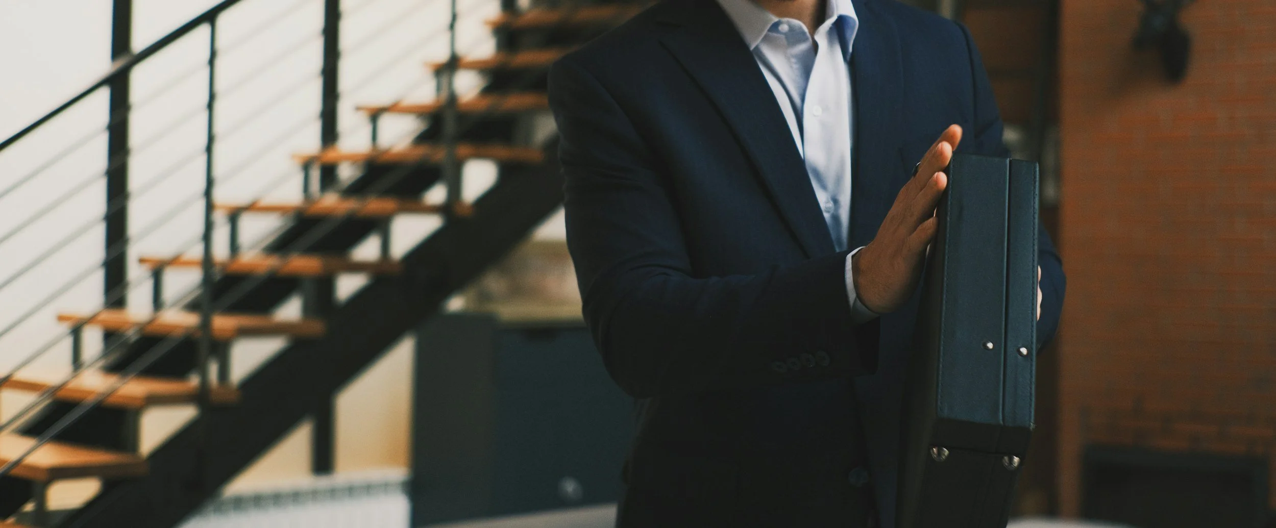 Un homme en costume tenant un porte-documents dans un intérieur moderne avec une échelle en métal et bois en arrière-plan.