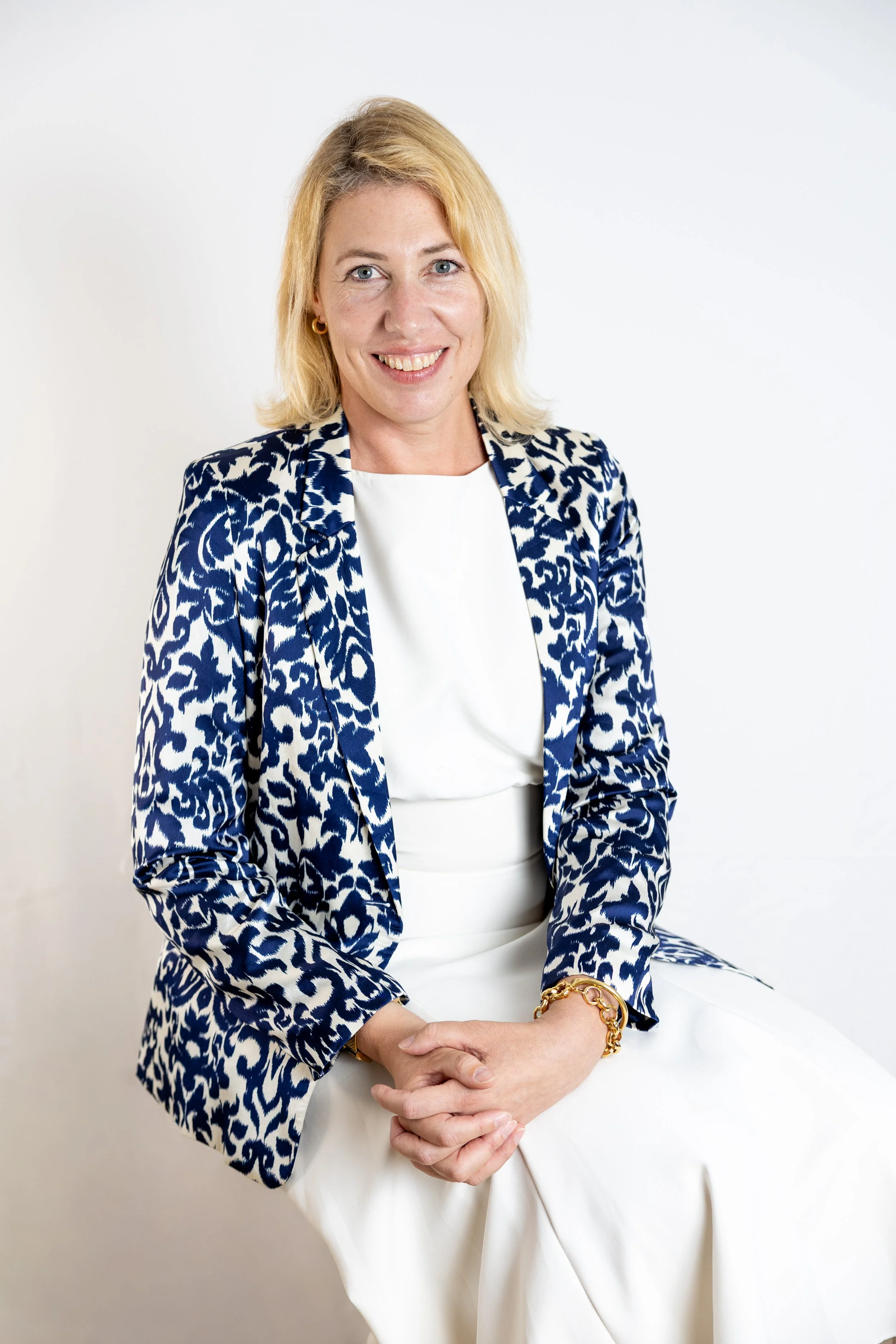 Une femme blonde souriante, vêtue d'un blazer à motif animal et d'une robe blanche, assise contre un mur blanc, portant des bijoux en or.