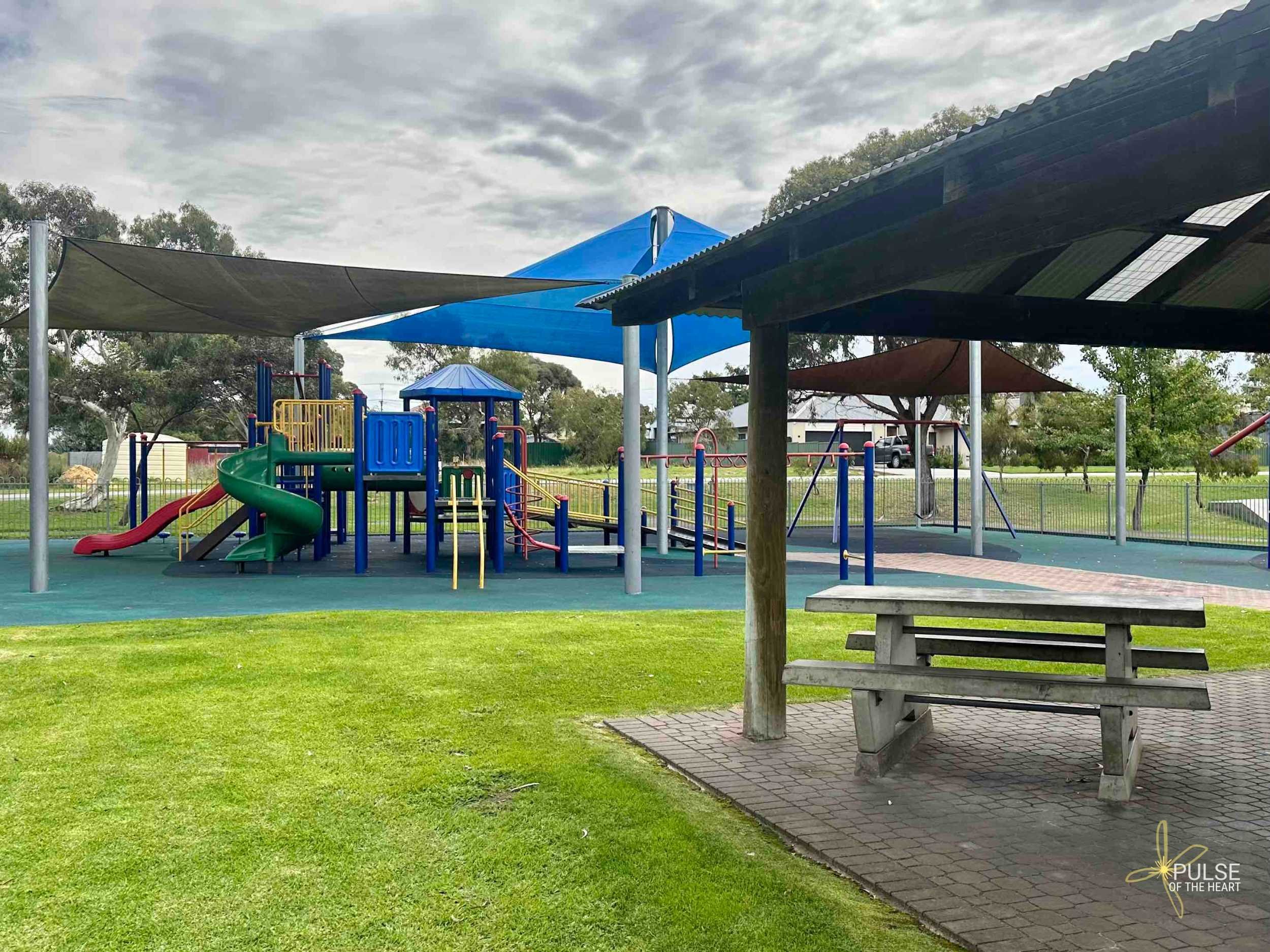Shaded picnic table and playground equipment at Market Square Recreation Area. Some softball, some grass.
