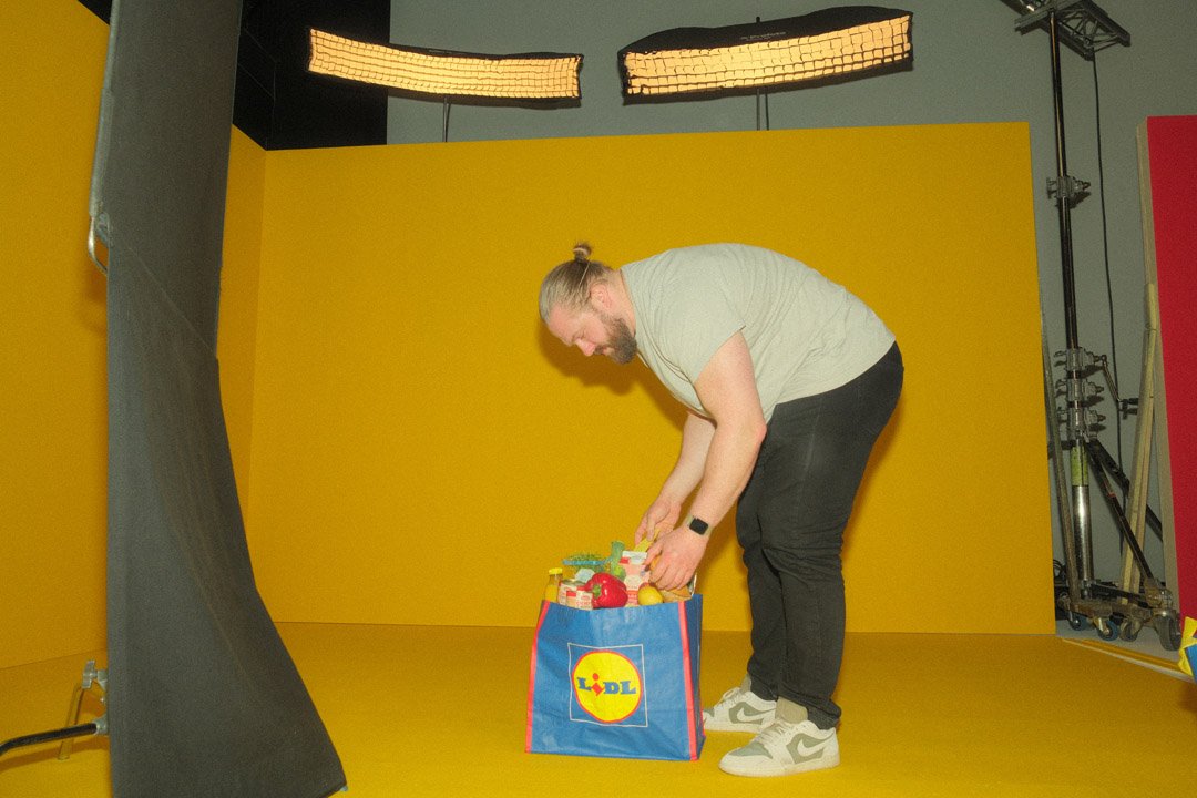 Man packing a blue Lidl shopping bag with toys or products in a photography studio with yellow background.