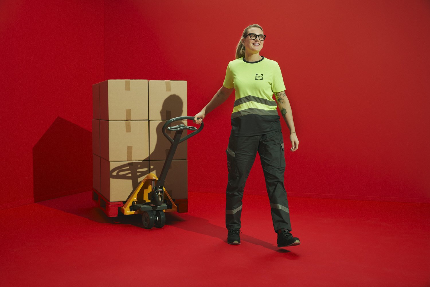 A woman in a lime green shirt and black pants with reflective strips stands against a red background, holding a pallet jack with stacked cardboard boxes.