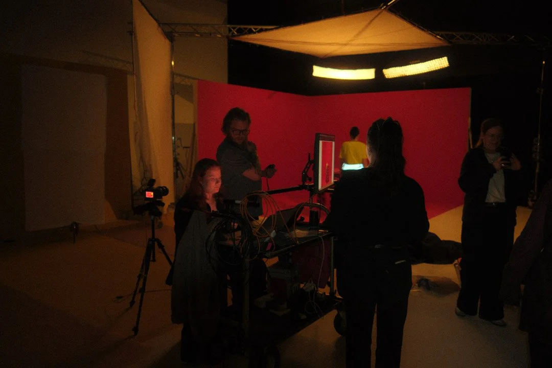 People working at computer stations with monitors, cameras, and lighting in a studio with a red backdrop and large overhead lights.