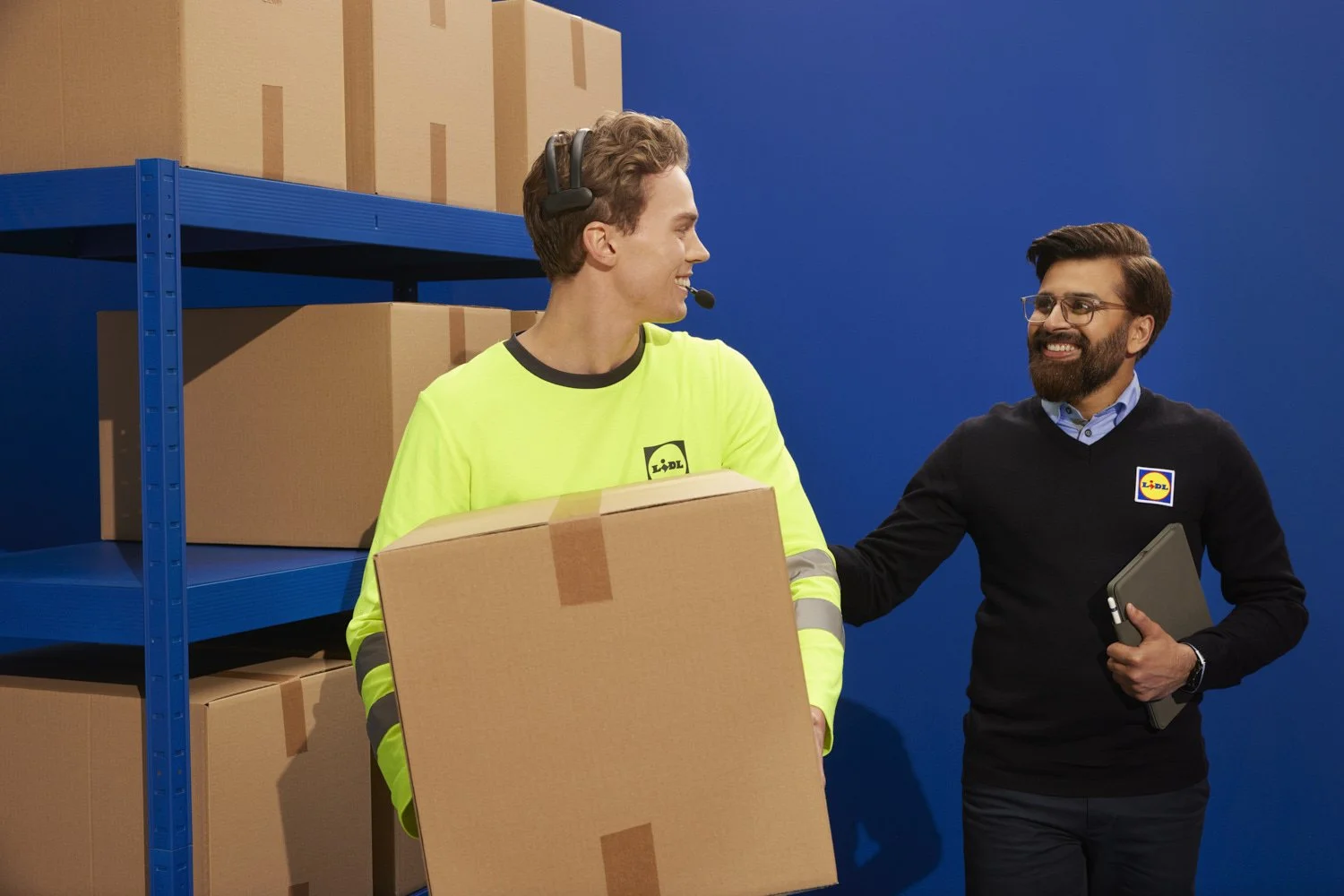 Two men at a warehouse, one in a neon yellow Lidl uniform, holding a box and wearing a headset, exchanging smiles with the other man who is in a black Lidl sweater, holding a tablet, in front of a blue wall and storage shelves with boxes.