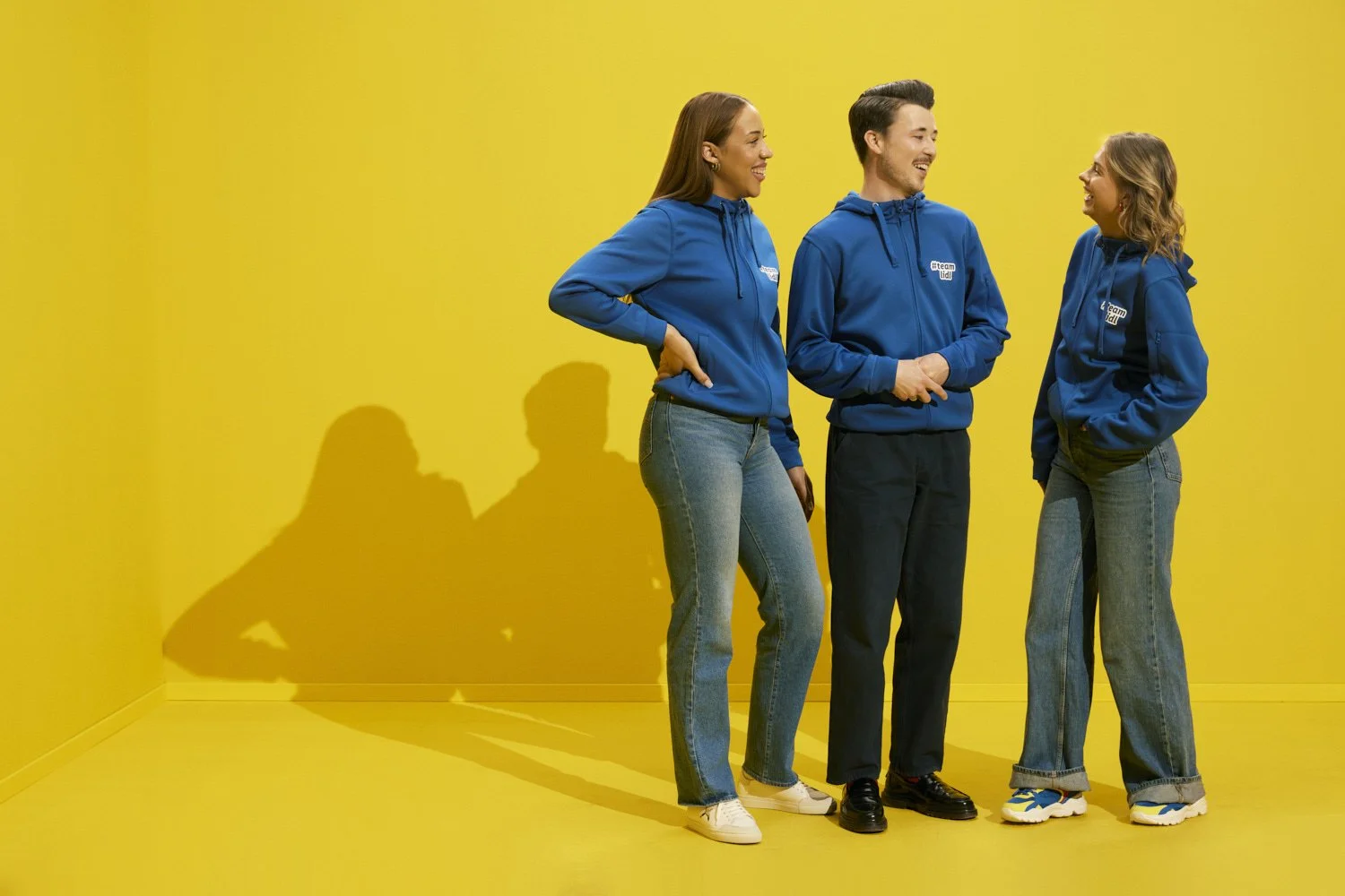 Three people, two women and one man, are standing and facing each other against a bright yellow background, wearing matching blue hoodies with a logo. They are smiling and appear to be engaged in a friendly conversation.