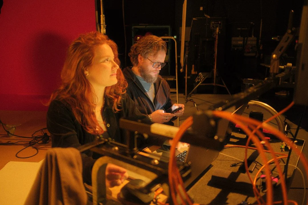 Two people sit at a table in a dark room with various electronic equipment and wires, one woman with red hair looking to the side and a man with glasses looking at his phone.