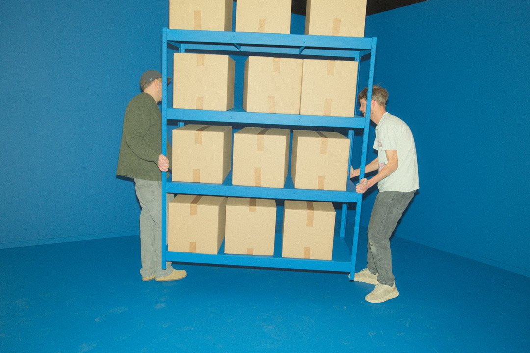Two men are moving a blue shelving unit filled with cardboard boxes in a room with blue walls and floor.