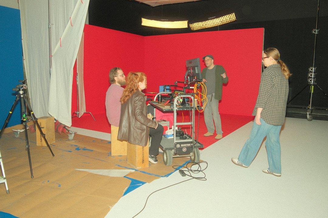 A group of five people on a video set with a red backdrop, filming equipment, and studio lights. One person is standing and talking to the group, while others sit or stand, with some seated on wooden blocks.