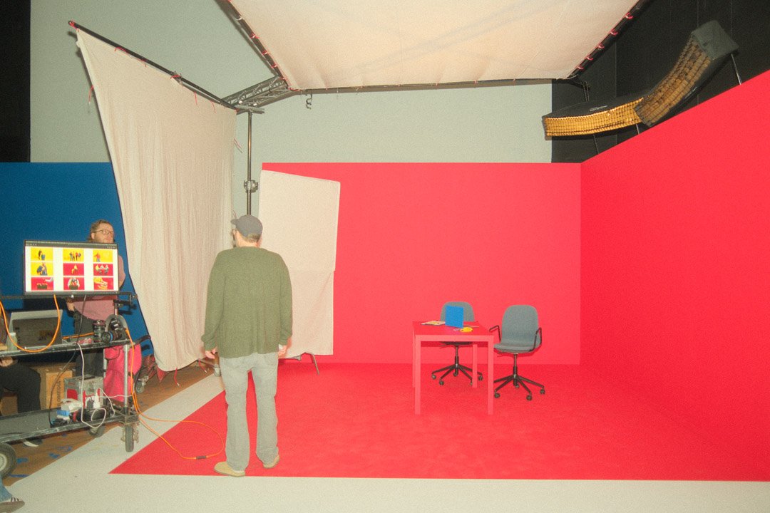 Photo of a photography studio with a red and gray backdrop, two chairs, a small red table with a blue chair, and technical equipment for filming or photography.