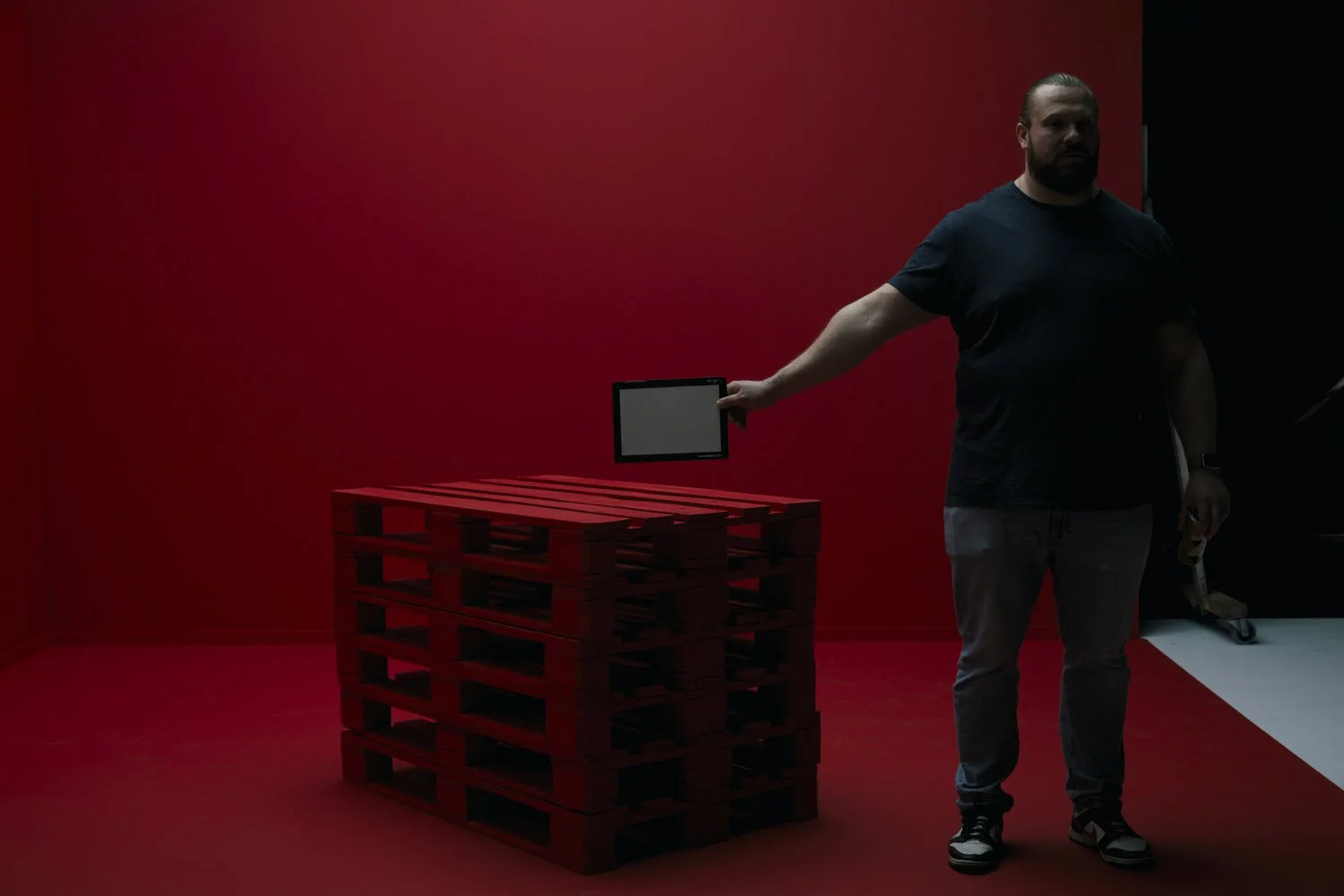 A man standing in front of a red and black background, holding a tablet in his right hand, with a stack of red pallets in front of him.