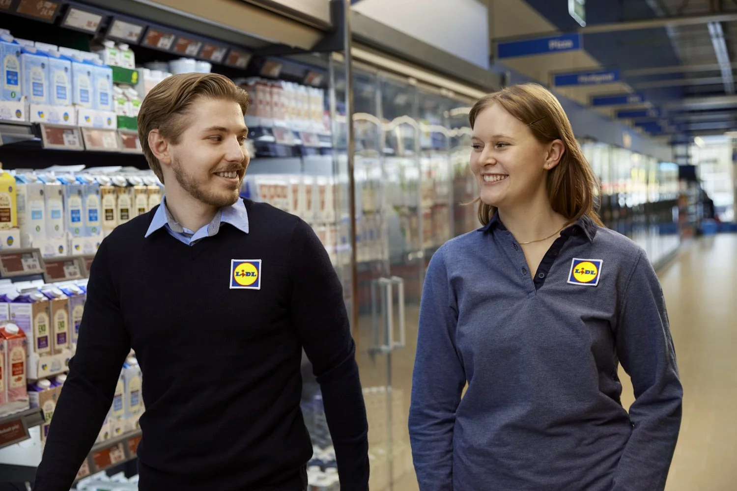 Two employees with Lidl uniforms shopping aisle, smiling and talking in a grocery store.