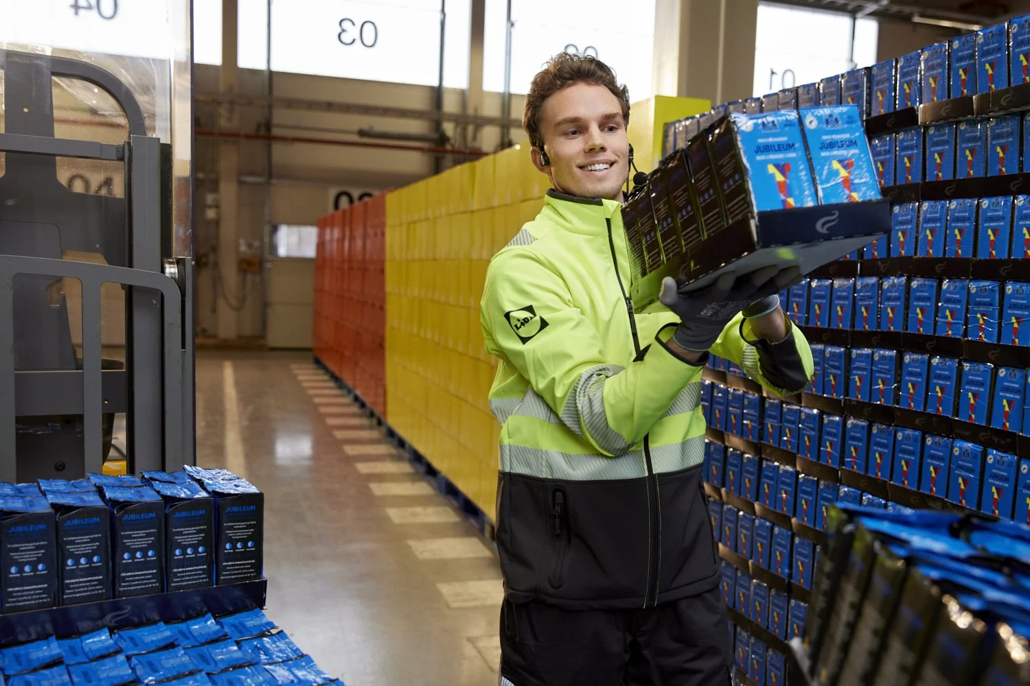 A man in a high-visibility jacket is managing products on shelves in a warehouse, with a background of colorful storage bins.